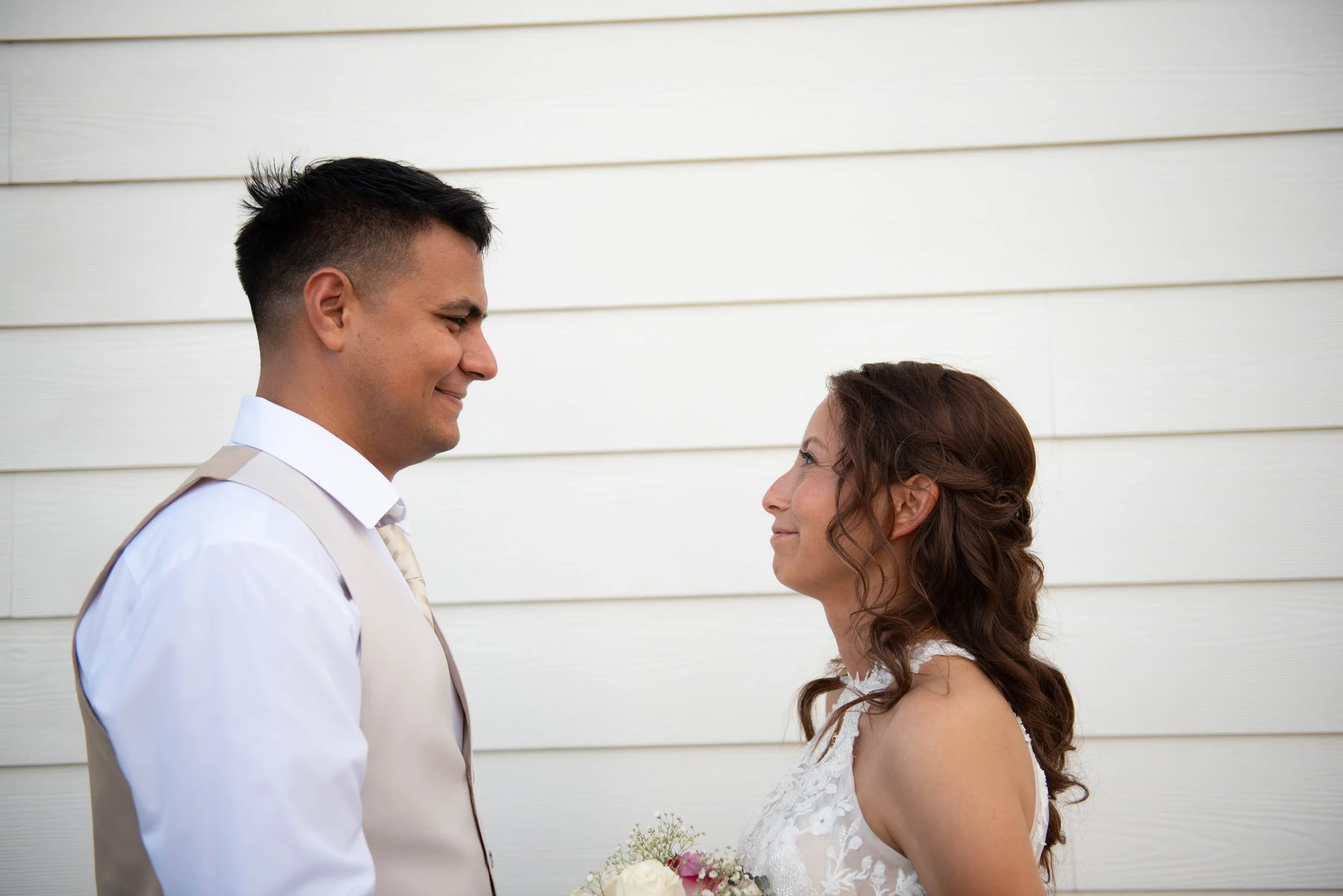 A bride and groom standing face to face, smiling at each other against a white wooden wall, during their wedding.