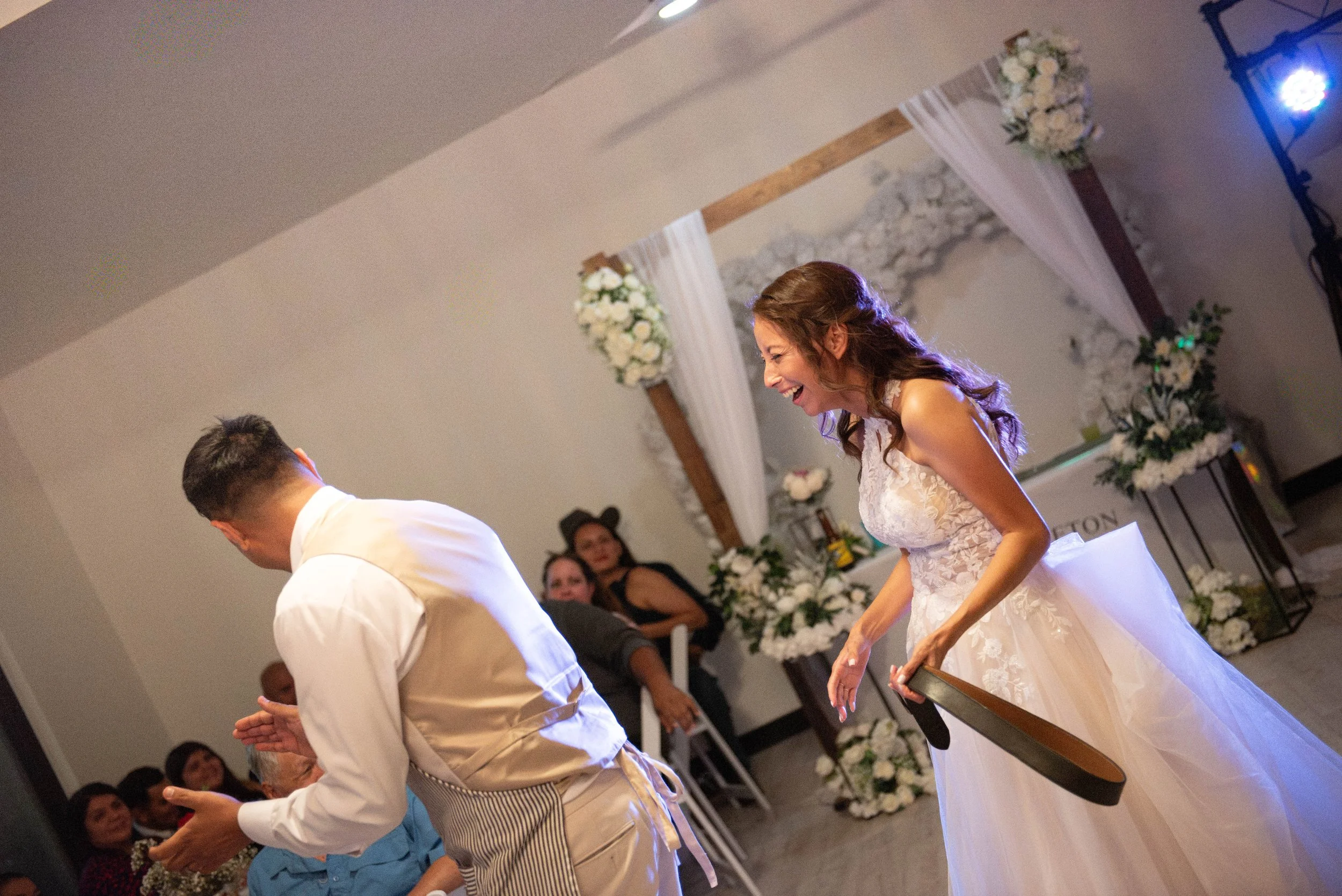 A woman in a wedding dress smiling during a wedding ceremony, with a man in a white shirt and apron. Several guests are seated in the background, and floral decorations with white flowers and drapes are visible.