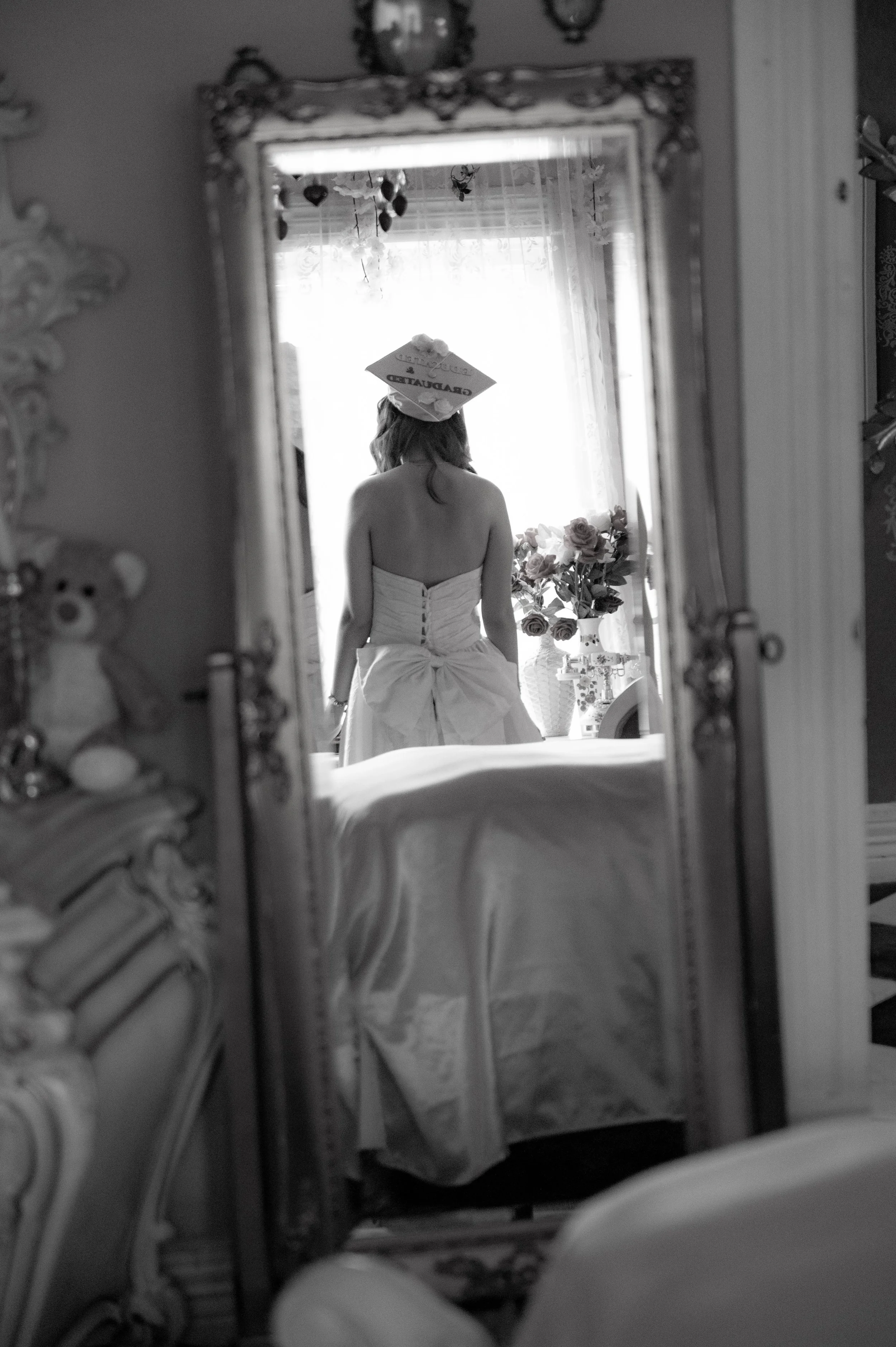 A young woman with long hair, dressed in a strapless gown, is seen from behind in a bridal or prom setting. She is wearing a decorated graduation cap. The photograph is taken through a decorative mirror, reflecting her back and a room with floral arr