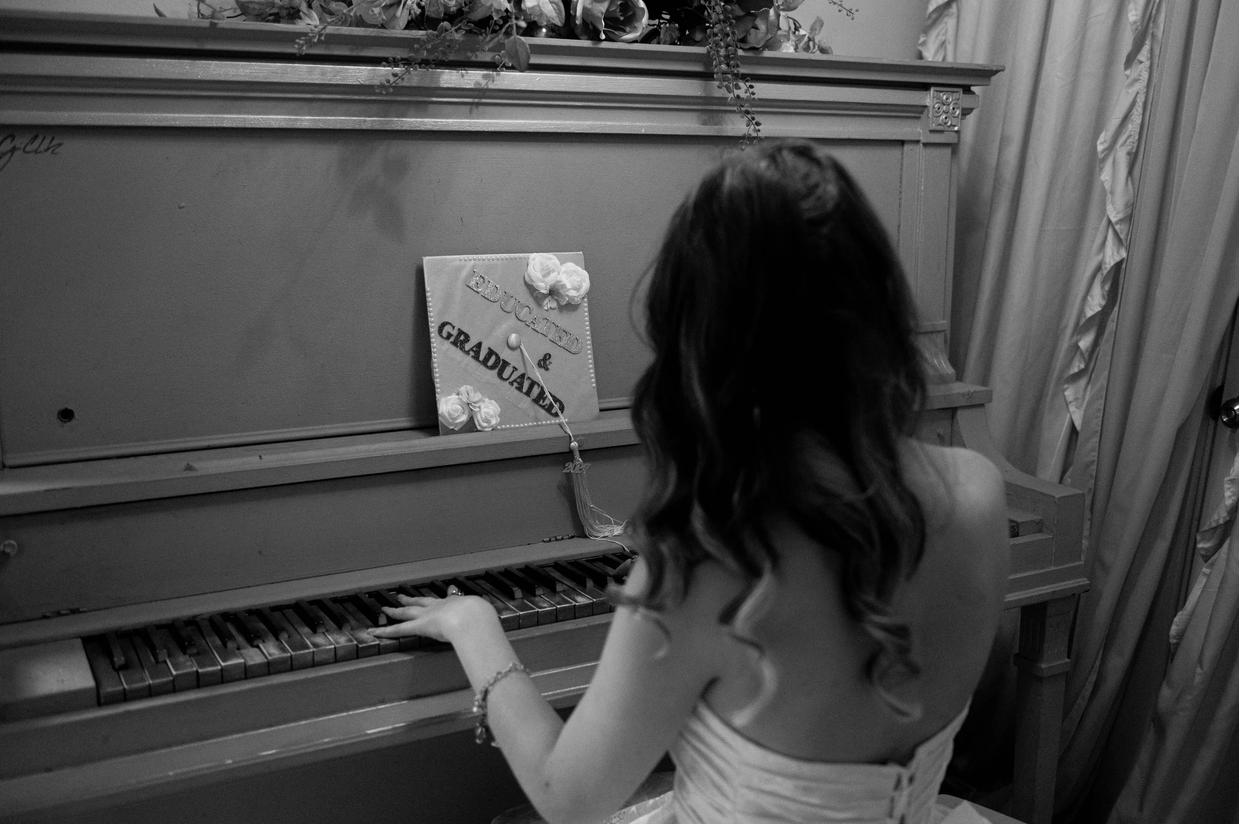 A young girl with long hair sitting at a piano, playing it. There is a festive sign decorated with flowers on the piano that says 'Education & Graduation.'