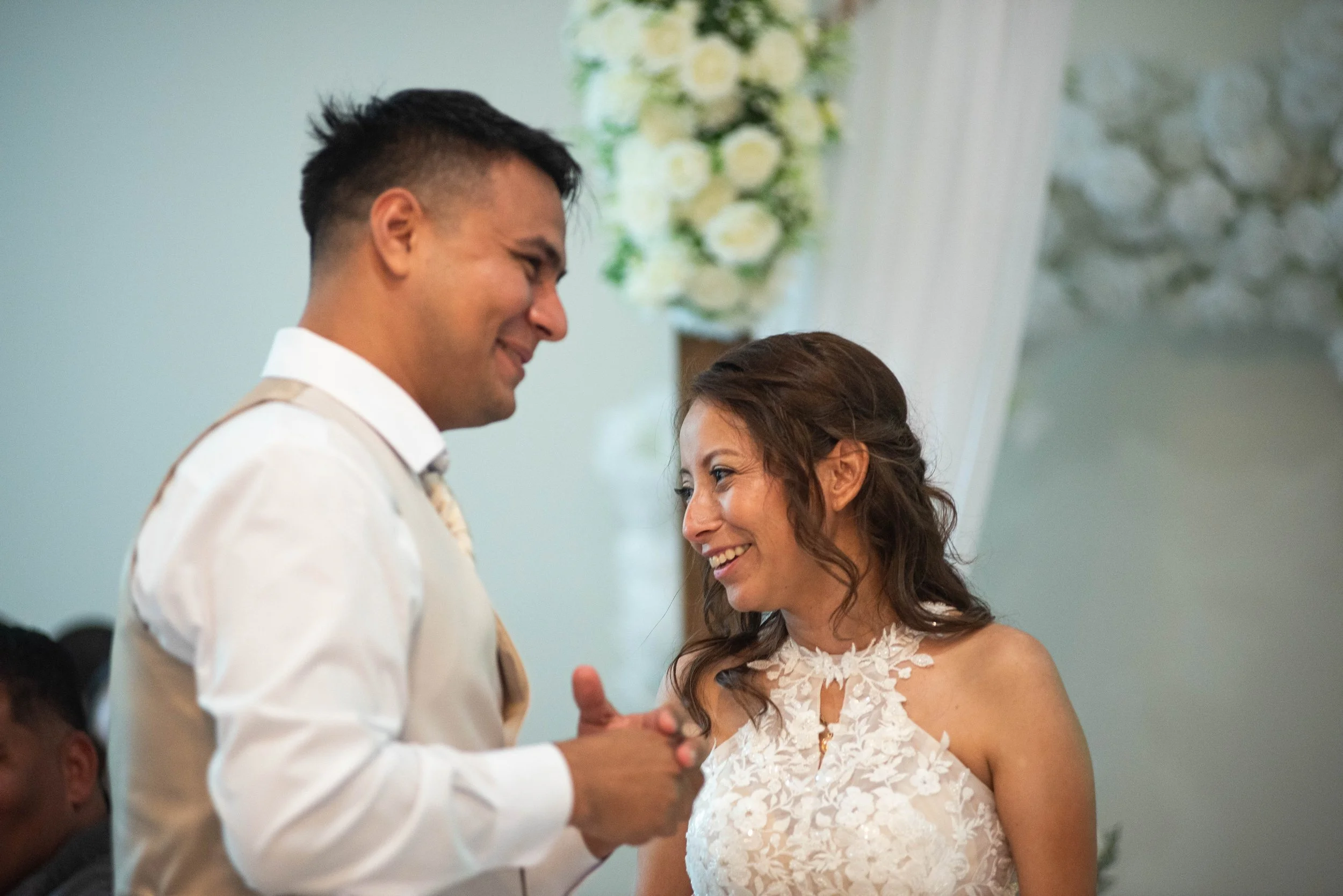 A couple sharing a joyful moment during their wedding ceremony, looking at each other with smiles, with a bouquet of white flowers in the background.