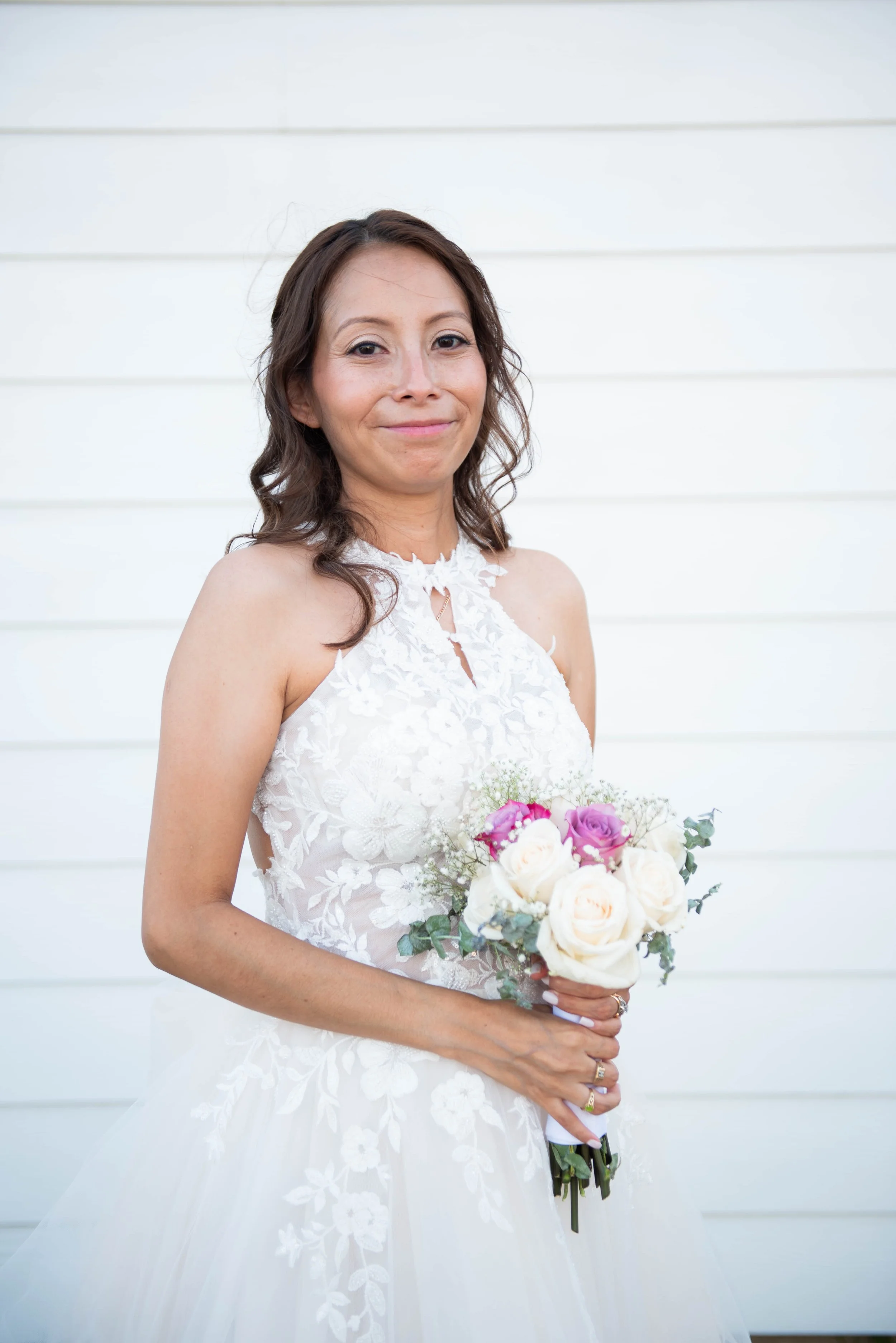 A woman in a white lace wedding dress holding a bouquet of white and pink roses, standing in front of a white wall.