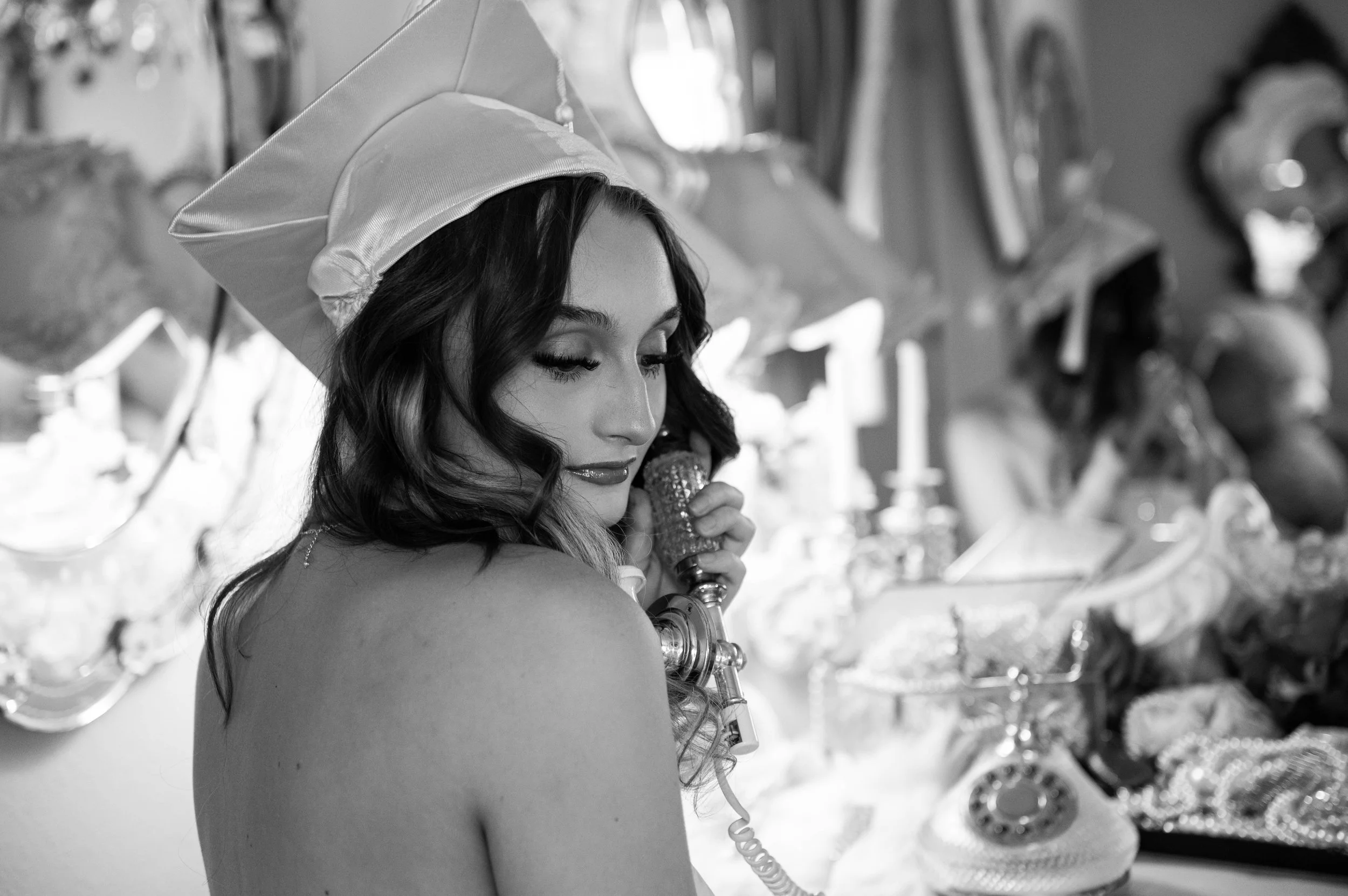 A woman with long wavy hair, wearing a graduation cap, looking down and holding a decorated bottle in her hand, with a row of shiny trophies in the background.