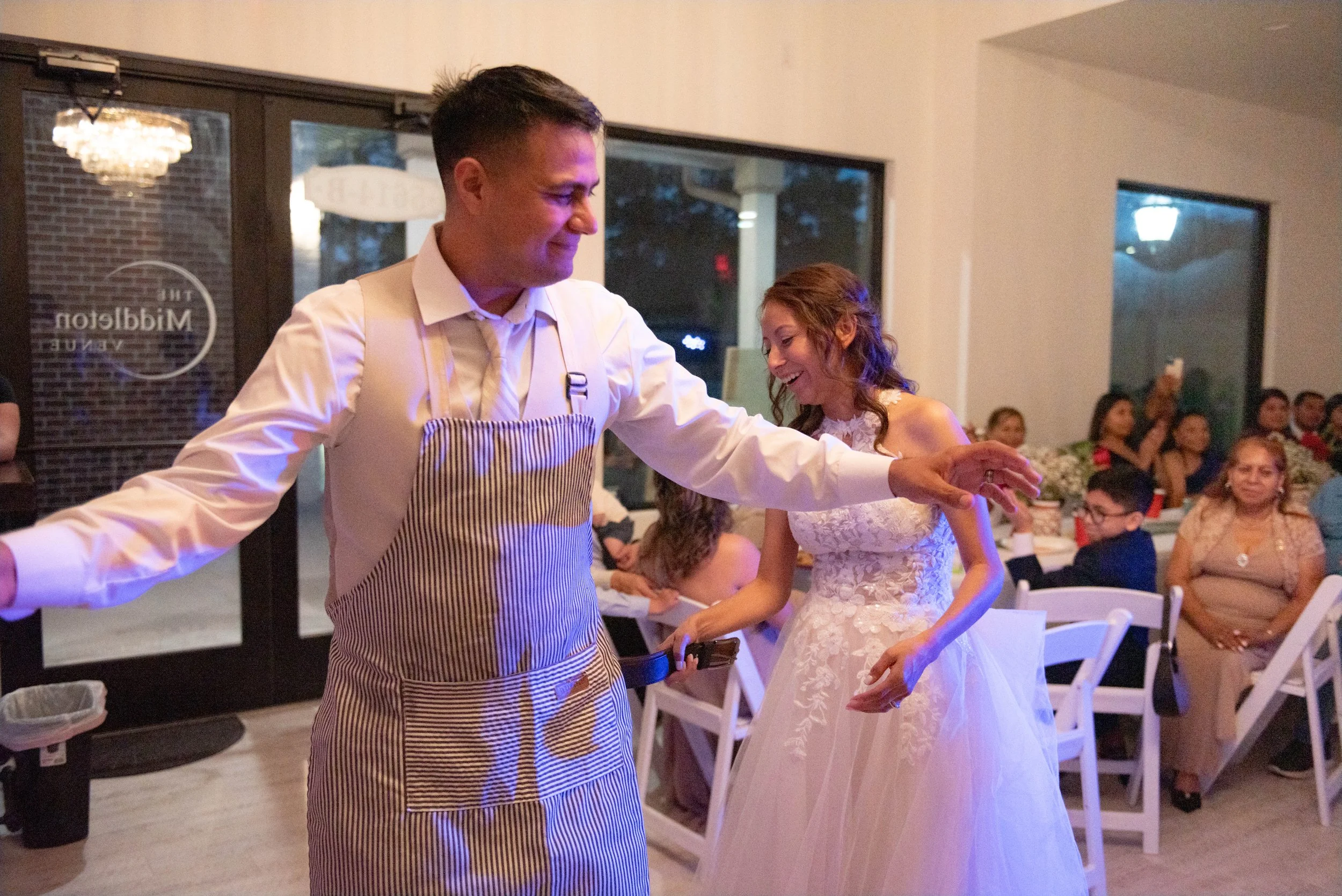 A man and a woman are dancing together at a wedding reception, with the woman in a white lace dress and the man in a white shirt and striped apron. In the background, guests are seated and watching, with some taking photos.