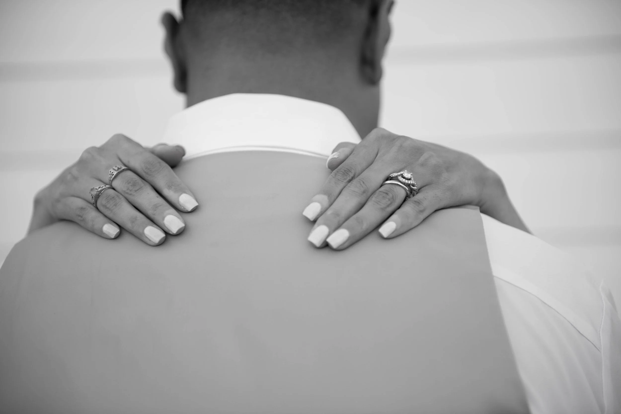 A person with rings on fingers is giving a hug to another person, seen from behind, in black and white.
