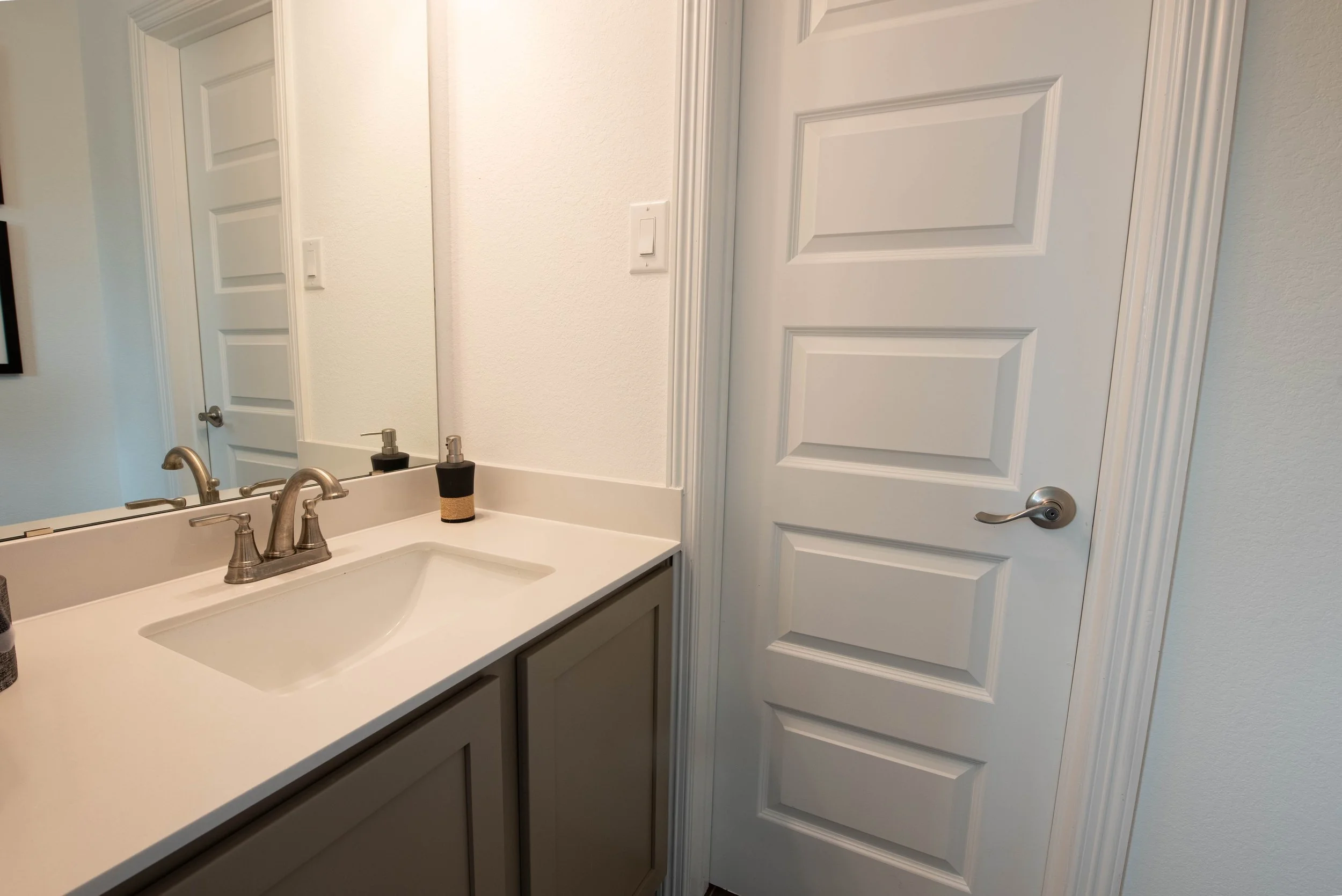 A bathroom with a white countertop, a silver faucet, a soap dispenser, a large mirror, and a closed white paneled door.