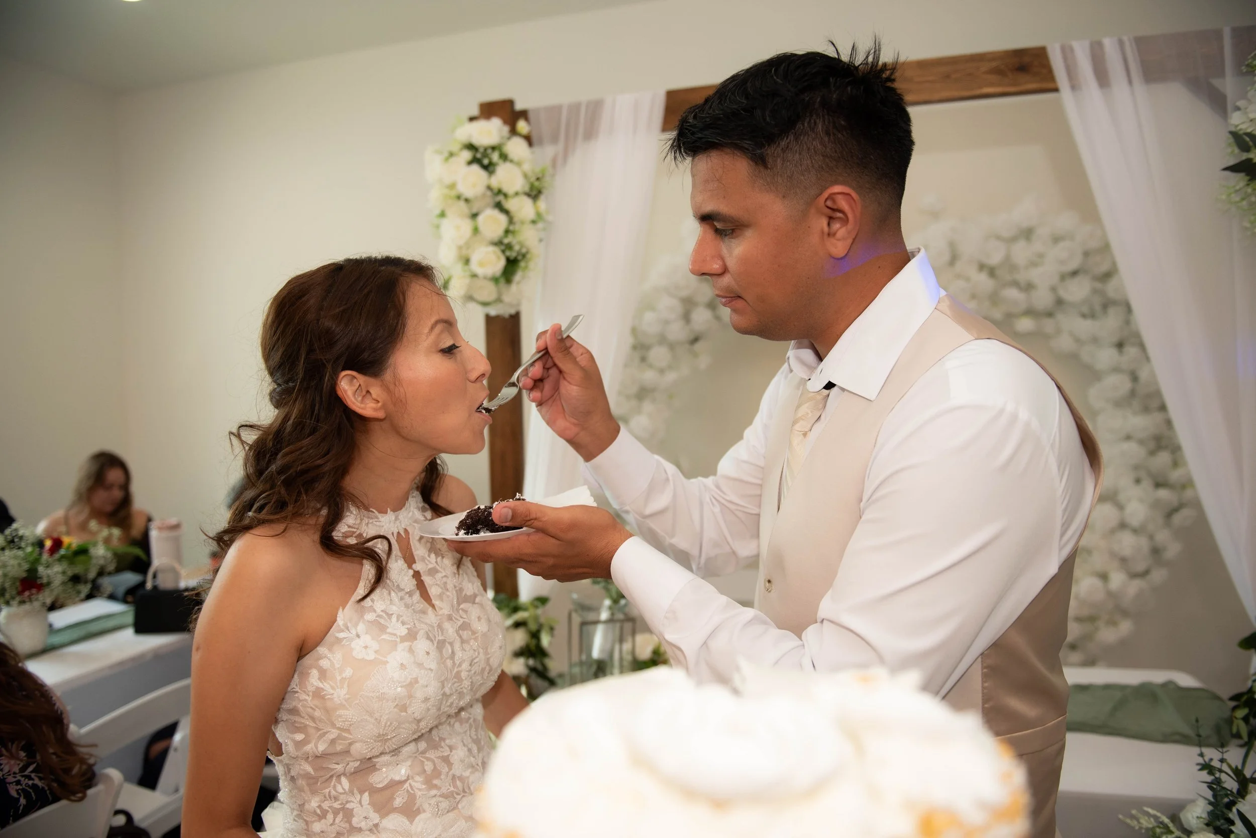 A man in a white vest feeds a woman in a white dress with cake during a wedding celebration in an indoor venue decorated with white flowers and drapery.