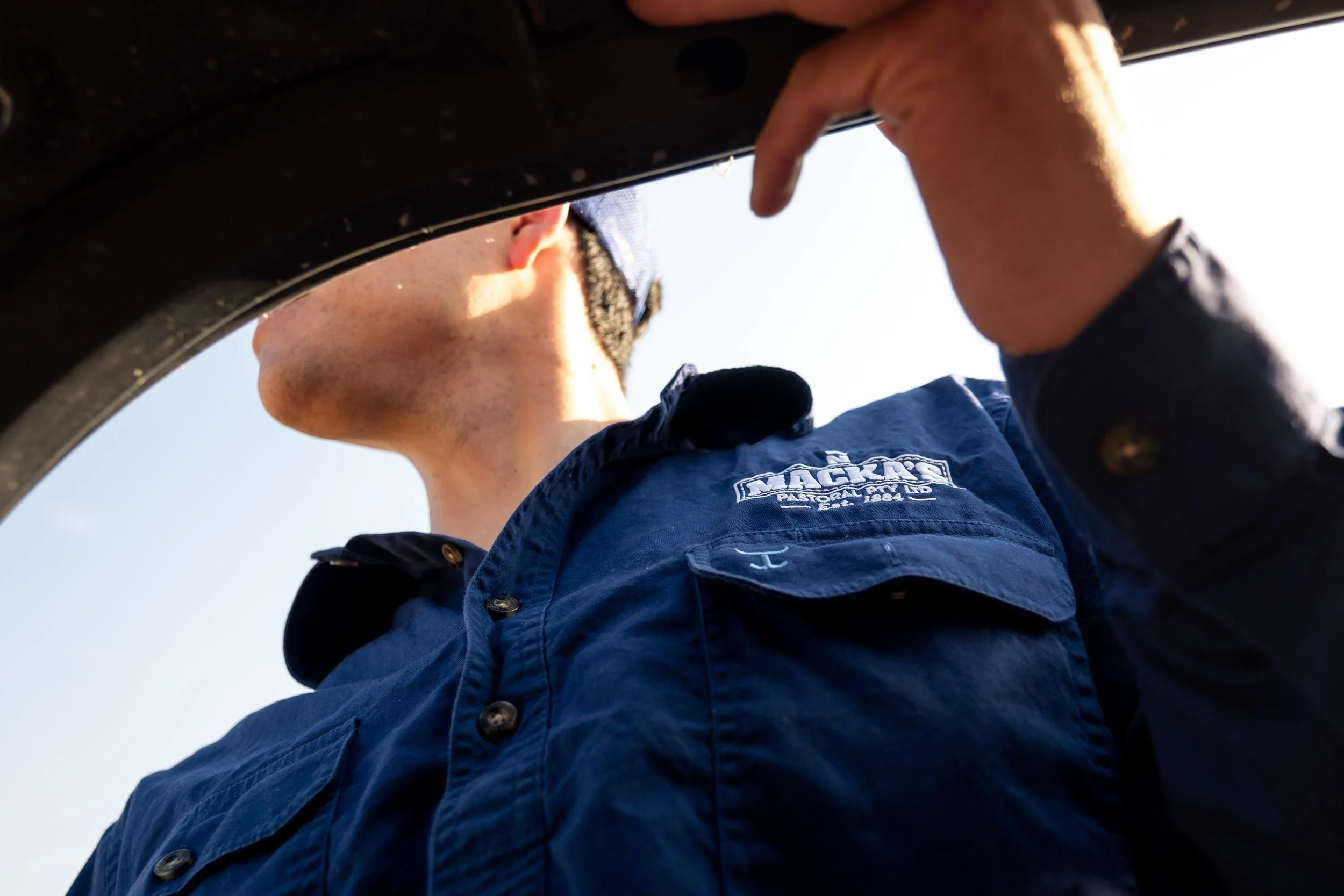 A young man wearing a blue uniform and a cap is looking down at the camera from below, with his hand near his face. The sky is visible in the background.