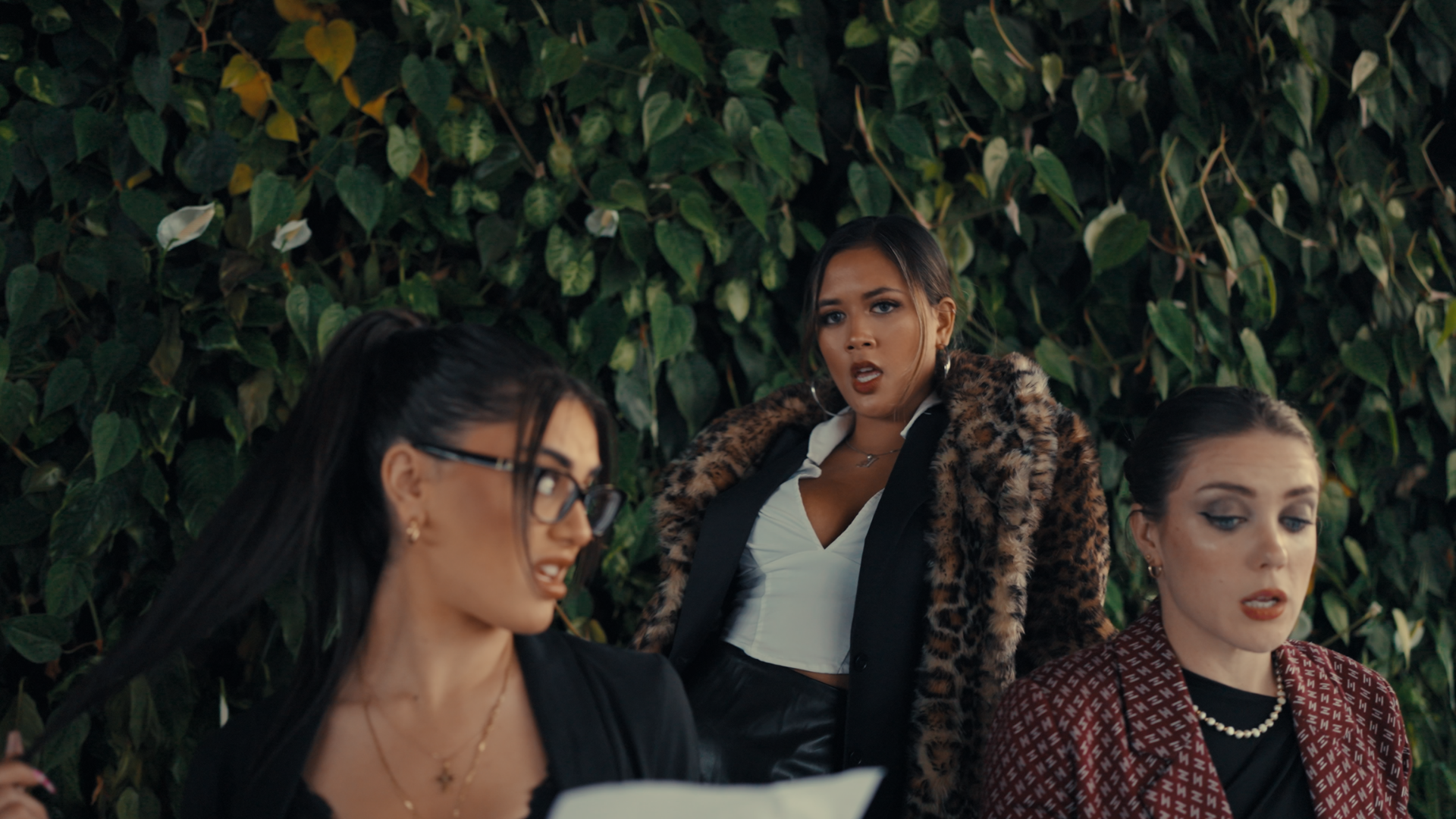 Three women sitting and standing in front of a green leafy hedge. The woman in the middle is wearing a leopard print fur coat and has an expressive facial expression. The women on either side are looking downward.