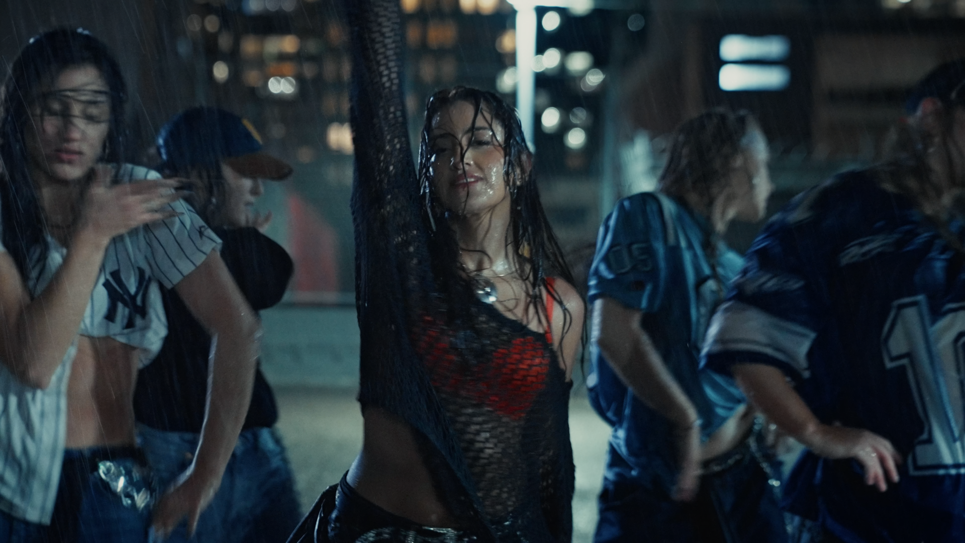 A woman with wet hair and a red and black outfit standing among others in rain at night. Player music video Aanya Melbourne Music Video.