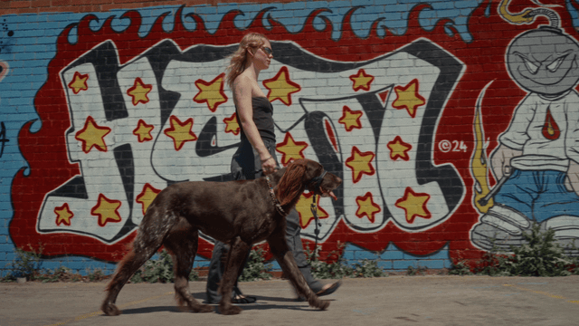 Woman walking large dog past colorful graffiti on brick wall.