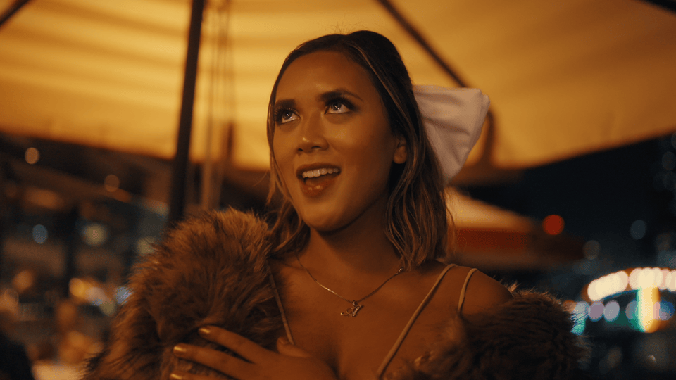 A young woman with shoulder-length hair, smiling and looking upwards, at night at an outdoor event with warm lighting. Melbourne-based R&B music video. Music video with Melbourne-based singer Alisya Rae. House Pop music. Melbourne city.