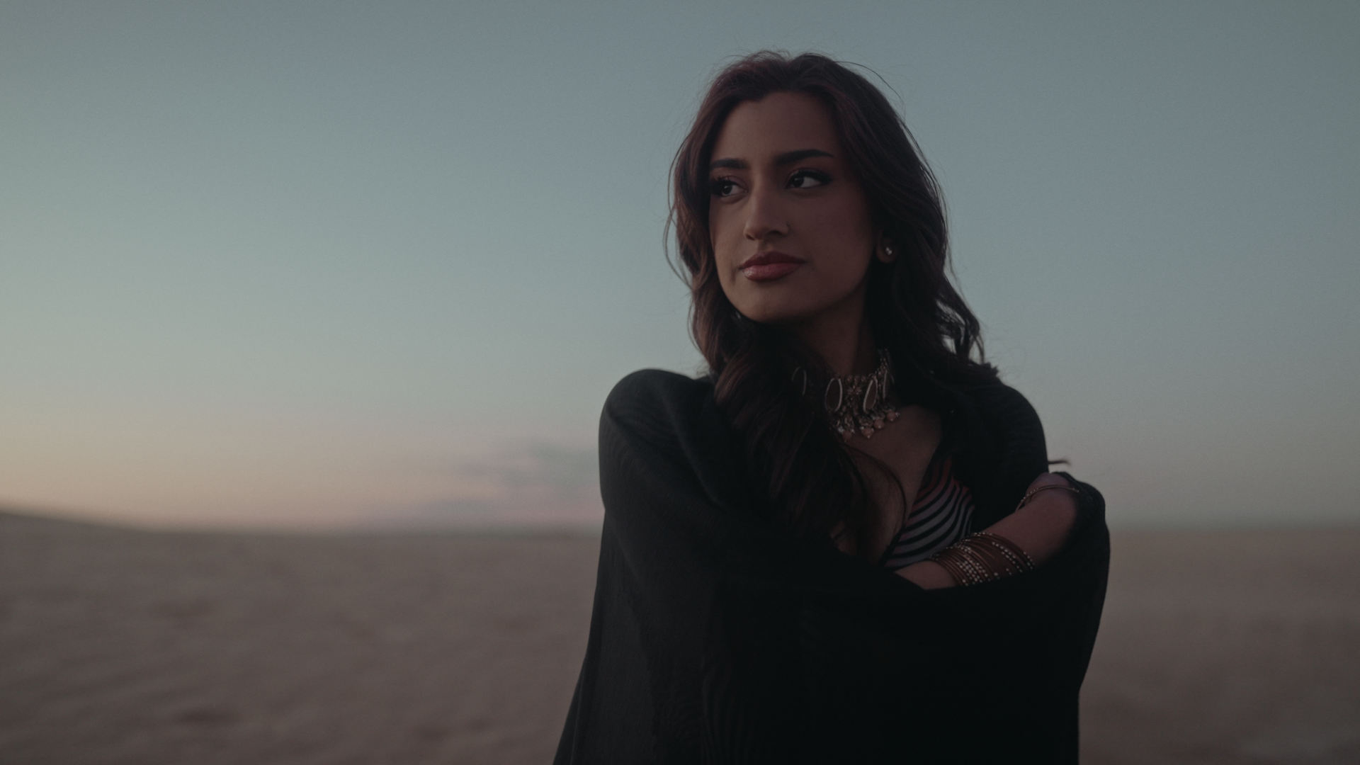 A woman with long dark hair and jewelry standing outdoors during sunset with a calm expression. Take Me Away music video Aanya Melbourne Music Video.