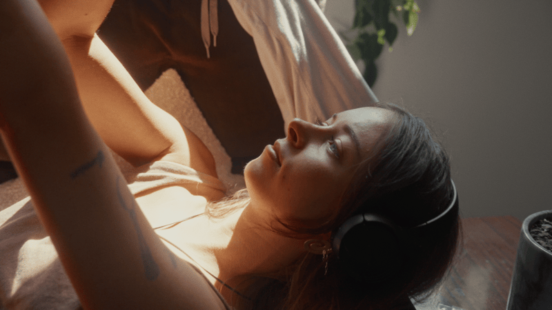 A woman lying on a bed wearing headphones, looking up with a relaxed expression.