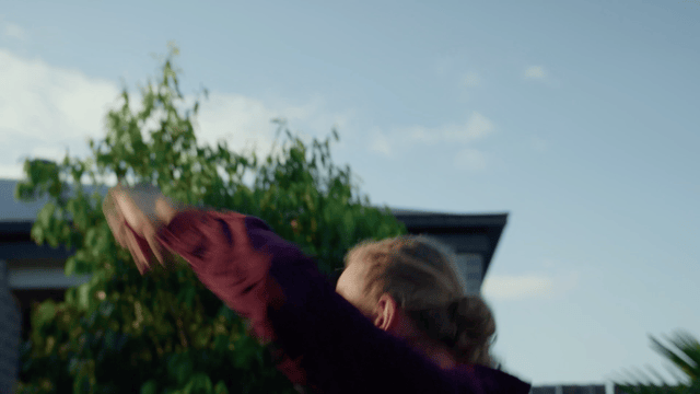 A person with long hair and a maroon jacket is outside, reaching upward with both arms, with green trees and a building in the background under a blue sky. Melbourne-based emotional real estate commercial campaign video. Melbourne production company.