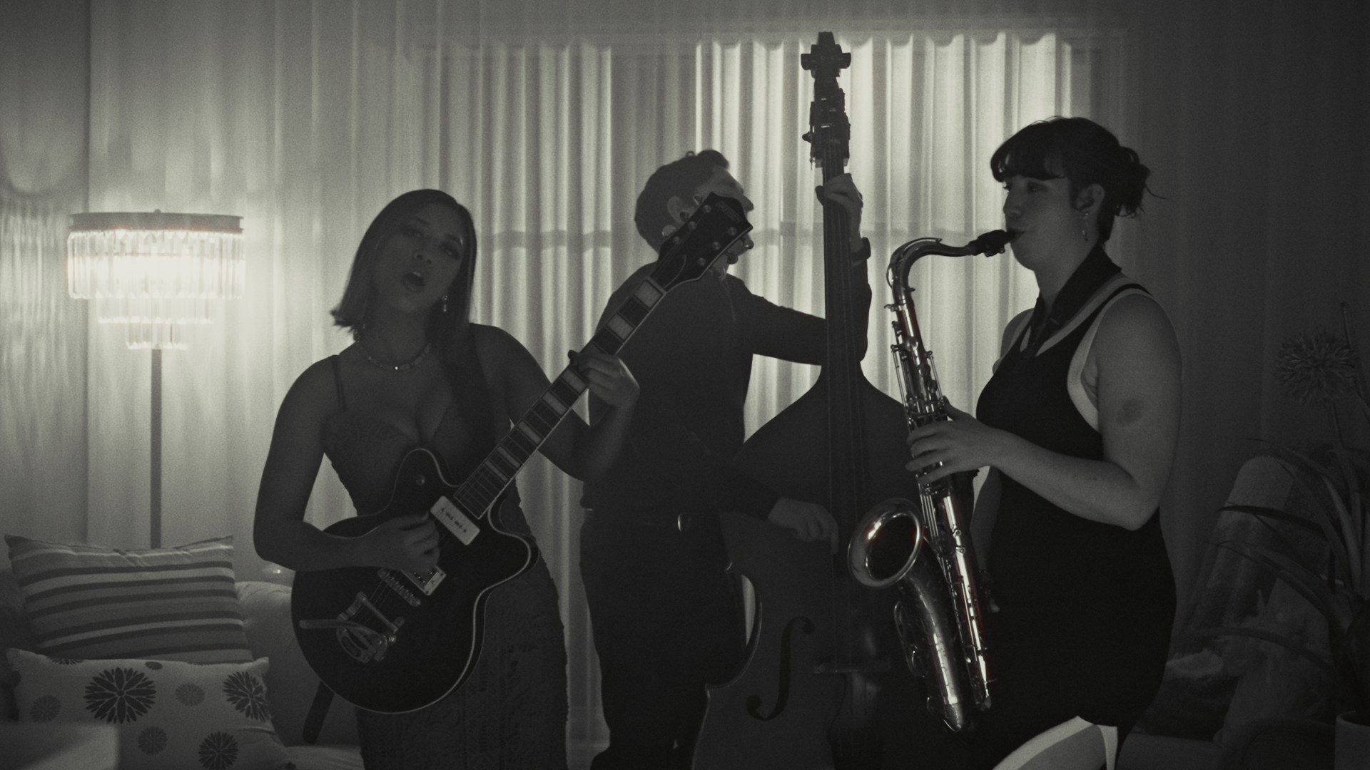 Three women playing musical instruments in a living room with a curtain backdrop. One woman is playing an electric guitar, another is playing an upright bass, and the third is playing a saxophone.