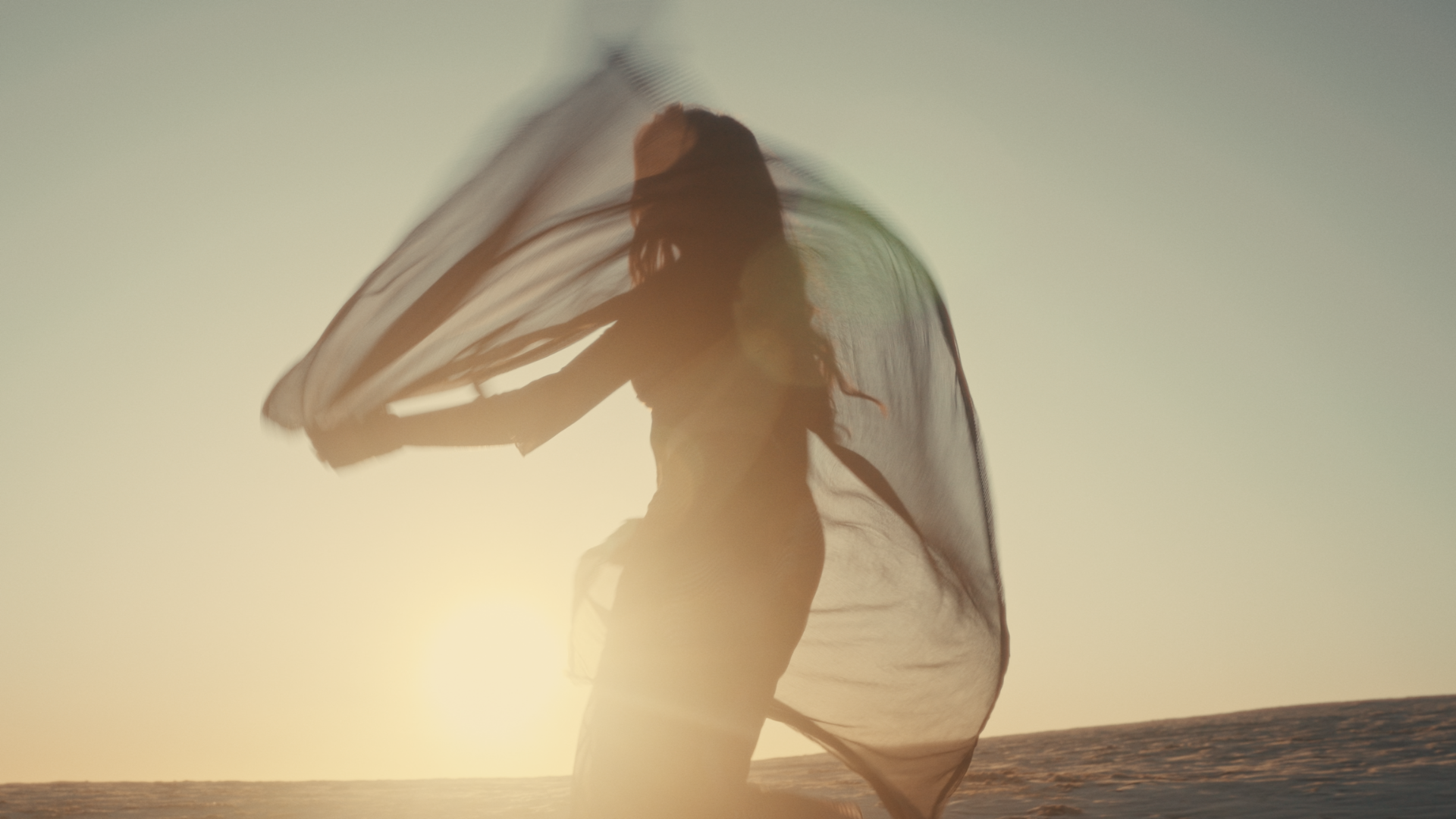 Silhouette of a woman holding a flowing sheer fabric near the beach at sunset. Take Me Away music video Aanya Melbourne Music Video.