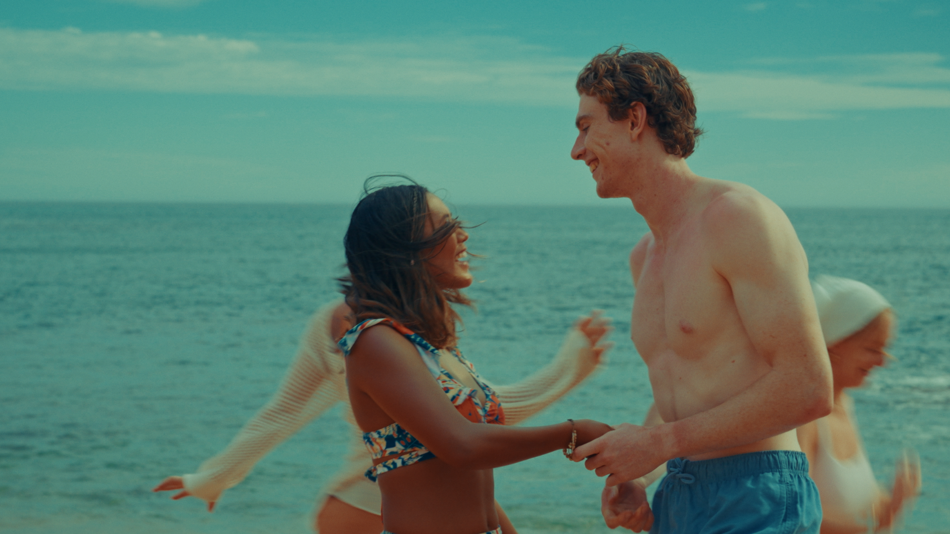 A young woman and a young man are smiling and holding hands on a beach, with the ocean and a cloudy sky in the background. Challengers-themed. Directed by Tea Van de Burgt.