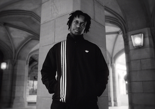 A young man with dreadlocks wearing a black jacket with white stripes and a small logo, standing against a stone pillar in an outdoor architectural setting.