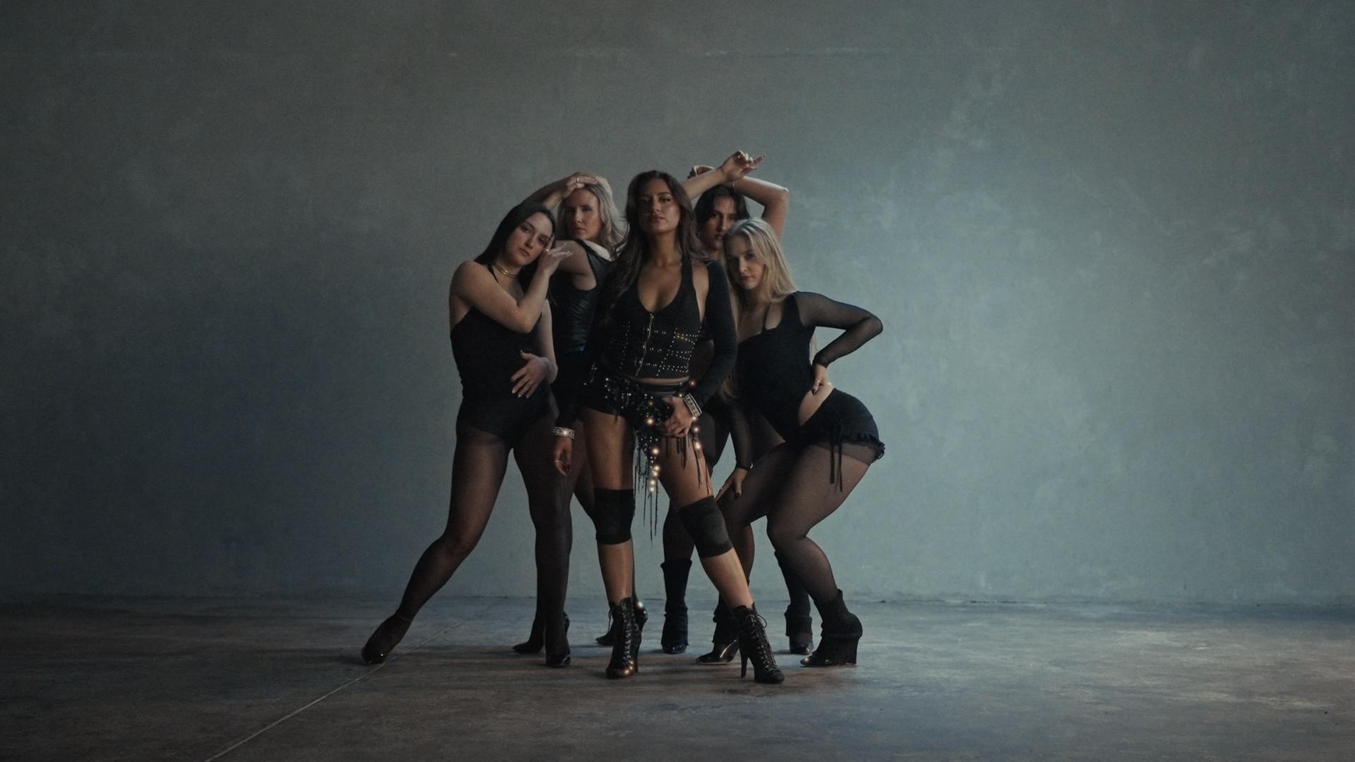 Group of five women dancing in a spacious, dimly lit studio with a plain, textured wall background. Player music video Aanya Melbourne Music Video.