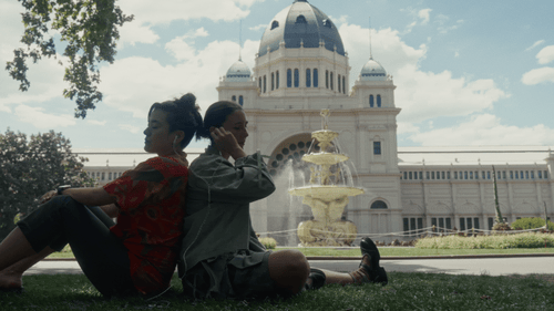 Two children sitting on grass near a fountain in front of a large, historic building with dome and multiple towers.