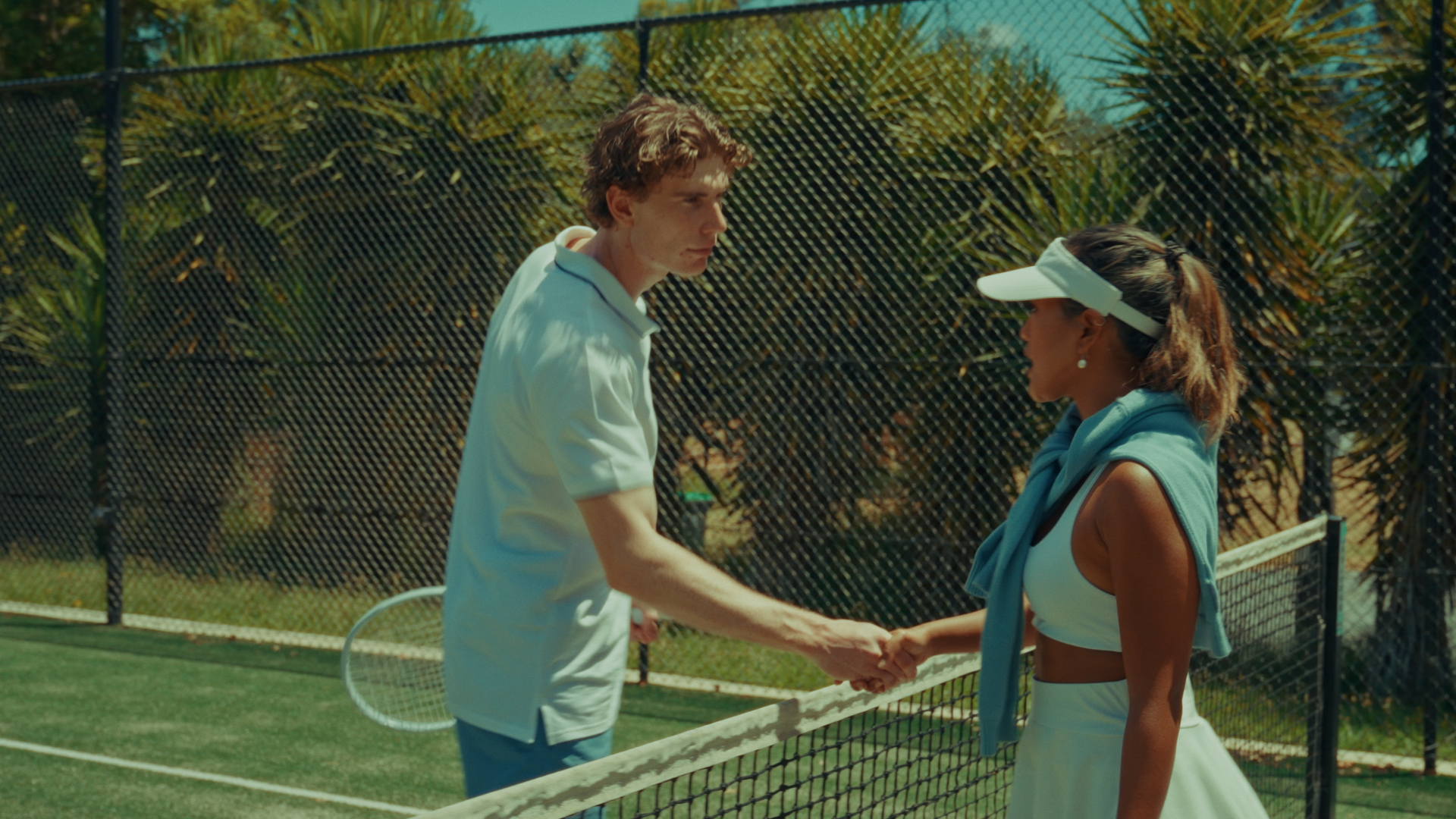 A man and a woman shaking hands on a tennis court with a net and green trees in the background. Alisya Rae 'Show Me' Music Video Melbourne. Challengers-themed. Directed by Tea Van de Burgt.