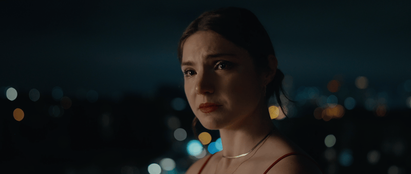 A woman with a somber expression standing outdoors at night against a blurred cityscape background with lights. Narrative short film, Beneath It All, Director Tea Van de Burgt, Cinematographer Darcy Michell. Drama emotional feminist short film