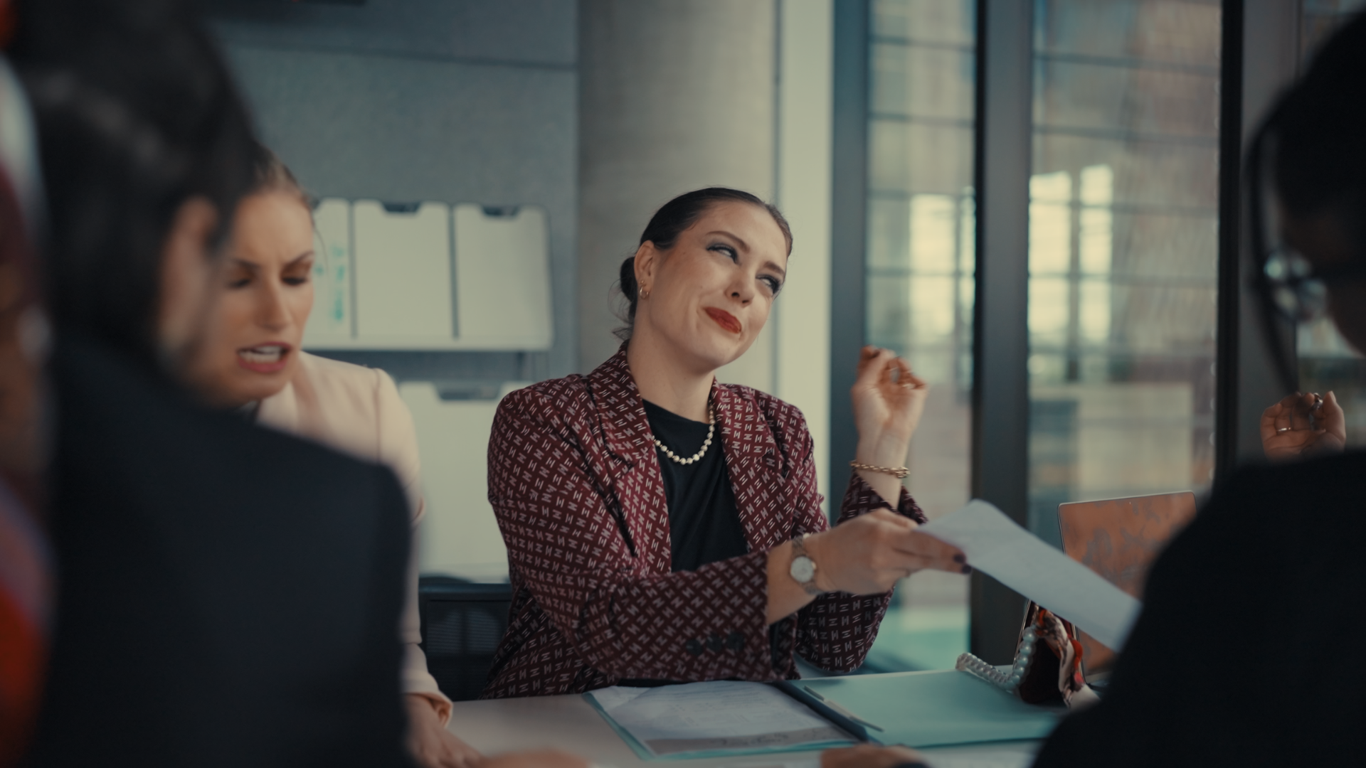 A woman in a red patterned jacket with a pearl necklace is sitting at a table with papers, talking to others in a business meeting. She is smiling and gesturing with her hand.
