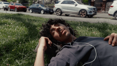 A young man with curly hair lying on grass listening to music with earphones outdoors near parked cars.