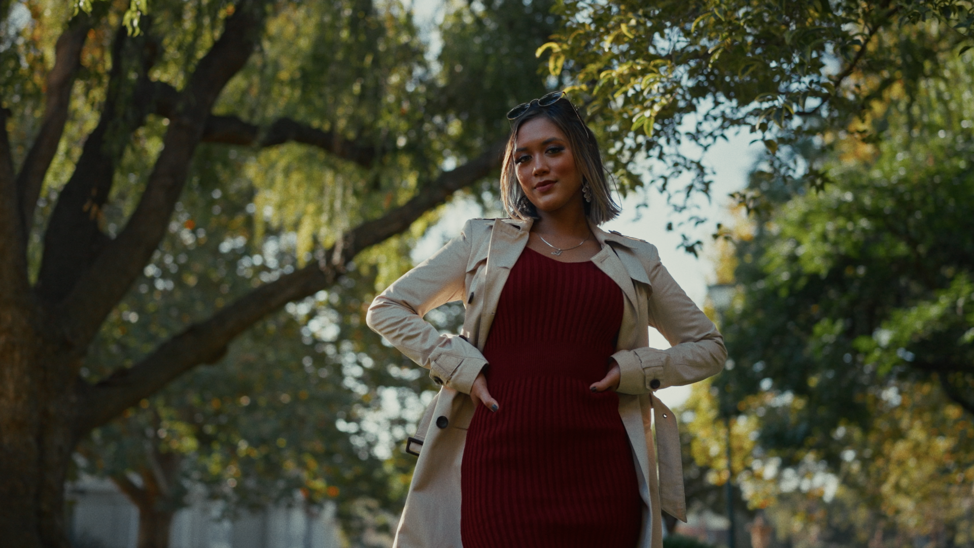 A young woman with dark hair standing outdoors in a park during autumn, wearing a beige trench coat over a red ribbed dress, with sunglasses on her head and jewelry, looking confidently at the camera.