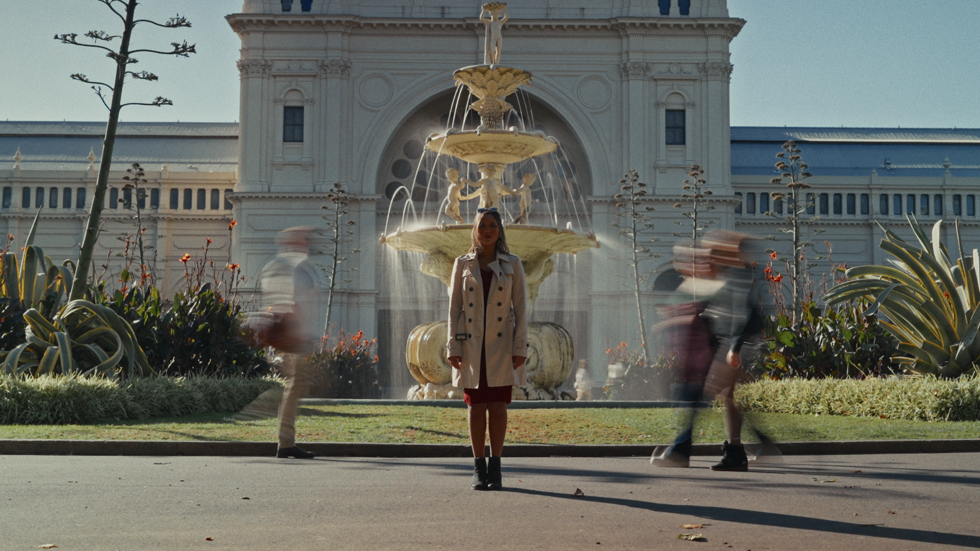 A woman in a beige coat stands in front of a multi-tiered ornate fountain with classical sculptures, behind a garden with plants, in front of a large, historic building. Several blurred people walk past in motion.