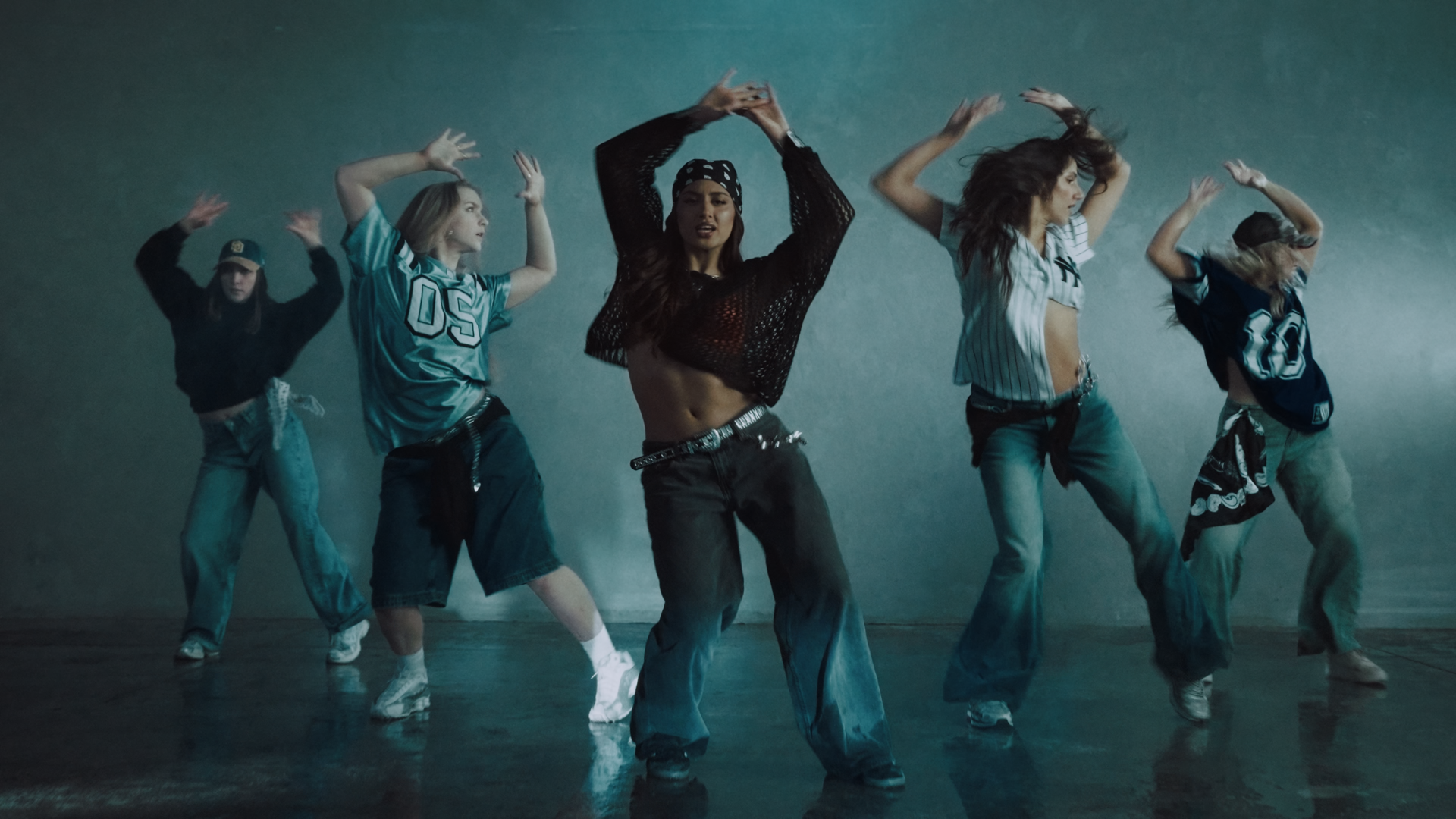 Group of young women dancing in a studio, dressed in casual streetwear and sports jerseys, with one woman in the center wearing a black mesh top and bandana, performing a dance move. Player music video Aanya Melbourne Music Video.