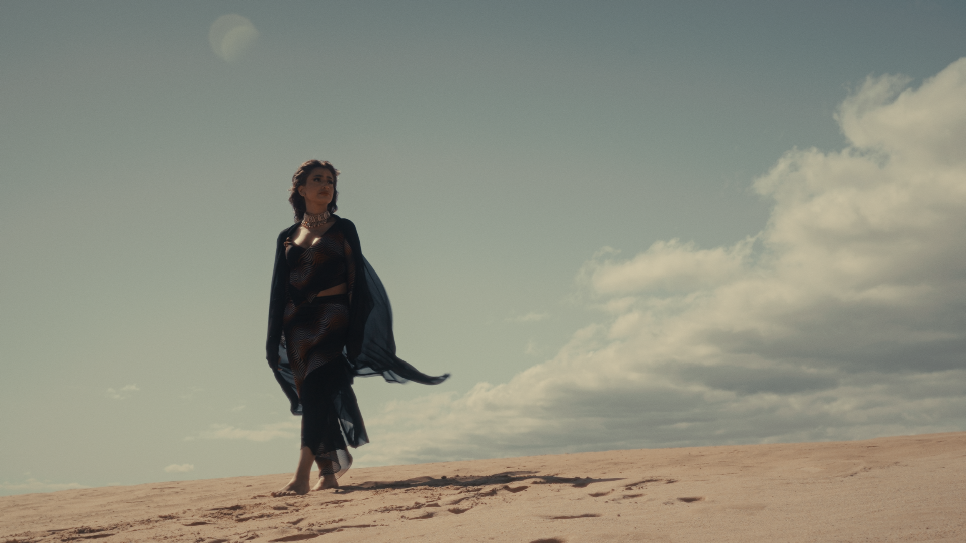 Woman walking on sandy terrain under a sky with clouds, wearing a black outfit with a sheer cover-up and jewelry. Take Me Away music video Aanya Melbourne Music Video.
