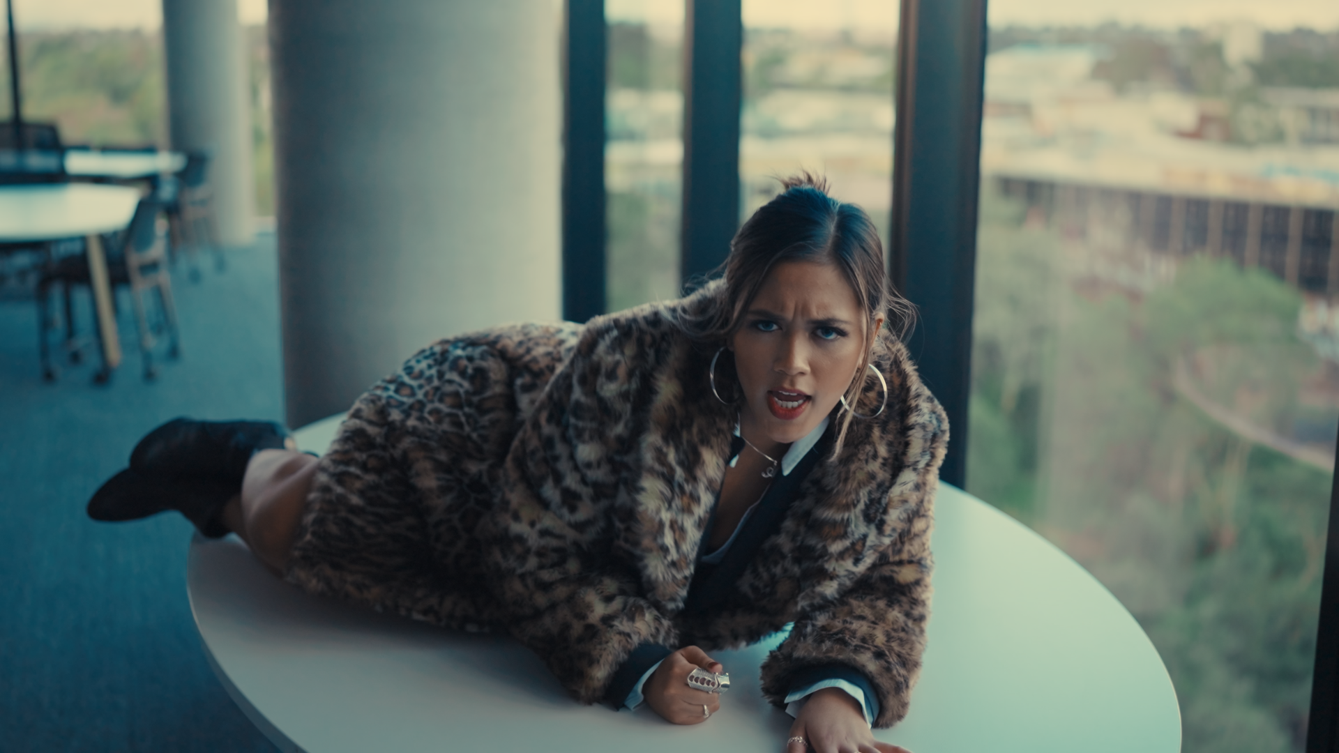 Woman with dark hair wearing hoop earrings and a leopard print coat, lying on a white table with a confused or angry expression, in a modern office with large windows and a city view.