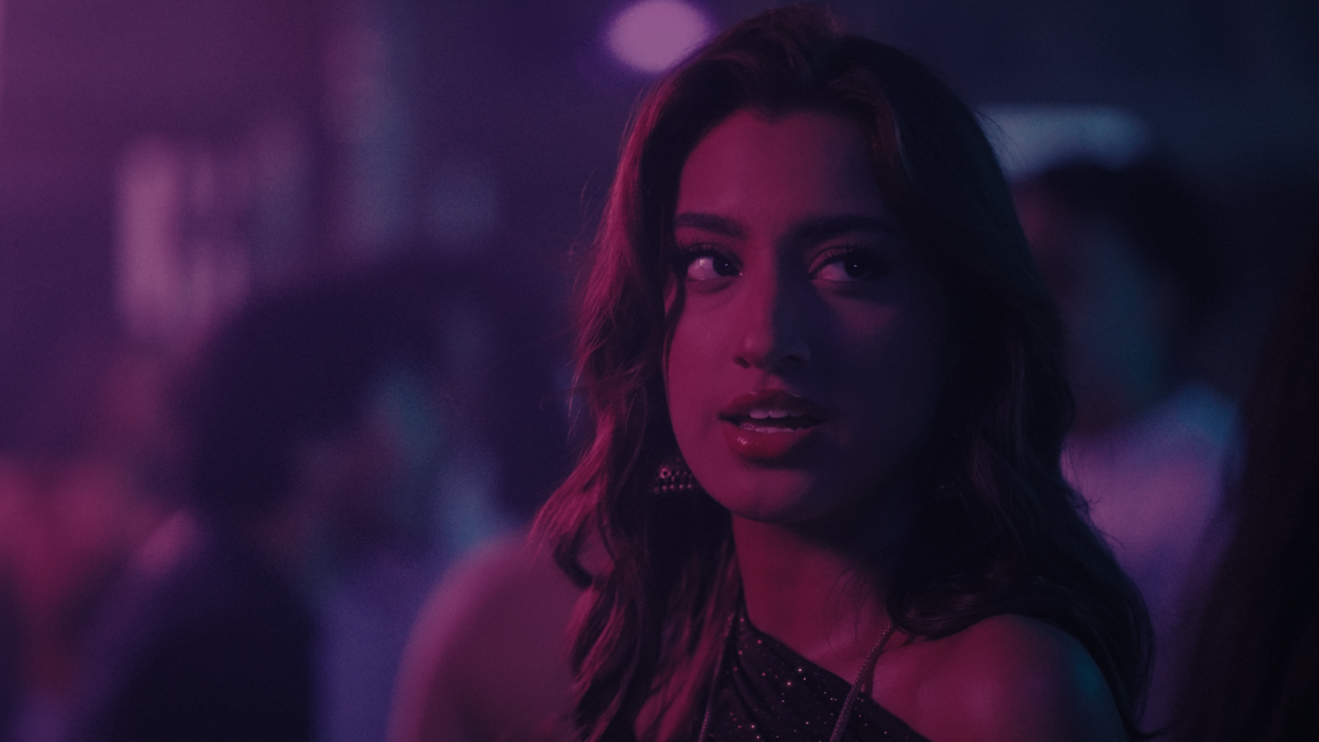 A woman with long, wavy hair and shiny lips looks over her shoulder in a dimly lit outdoor setting with purple and pink neon lights. Take Me Away music video Aanya Melbourne Music Video.