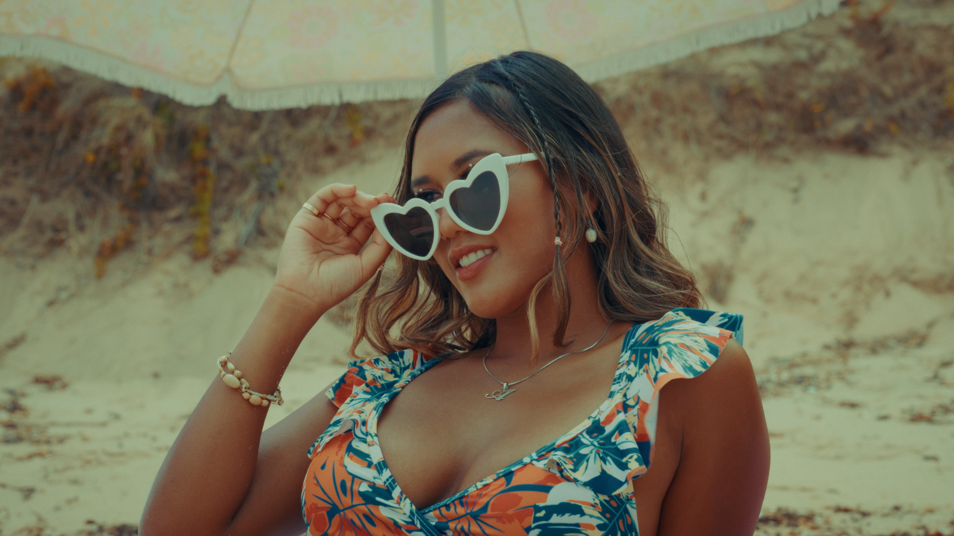 A woman wearing heart-shaped sunglasses and a floral dress stands on a beach, holding her sunglasses and smiling. Alisya Rae 'Show Me' Music Video Melbourne. Directed by Tea Van de Burgt.