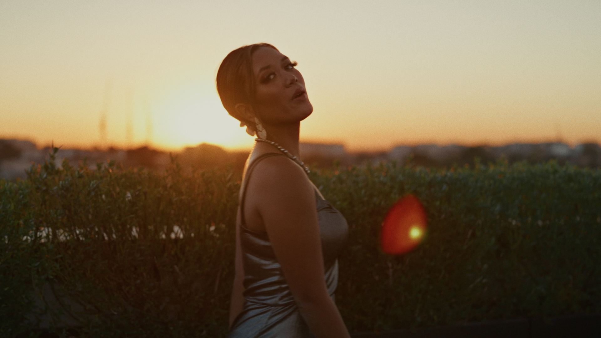 A woman with short hair standing outdoors during sunset, wearing a dress with thin straps, and a pearl necklace, with a hedge and cityscape in the background.