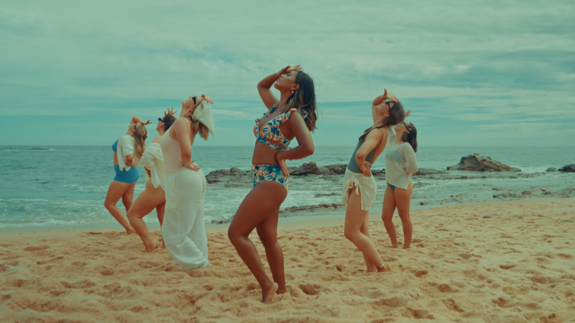 Group of women dancing on a sandy beach near the ocean, some with their eyes closed and hands on their foreheads, during daytime with cloudy sky. Alisya Rae 'Show Me' Music Video Melbourne. Directed by Tea Van de Burgt.