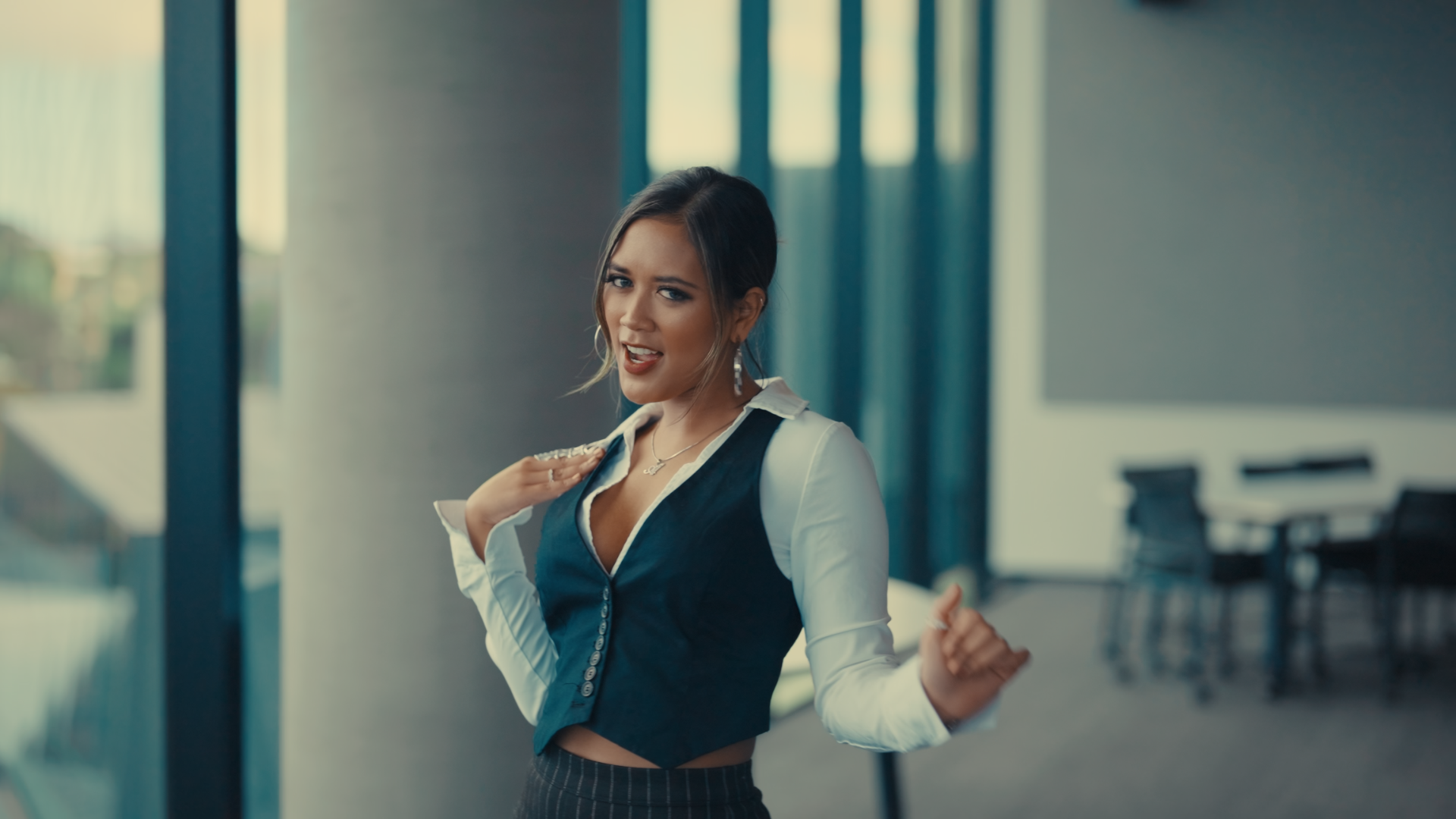 Young woman with dark hair styled in a loose updo, wearing a white blouse under a black vest, smiling, posing with a hand on her shoulder at an indoor location with large windows and chairs in the background.