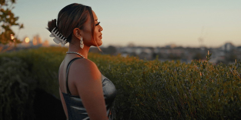 A woman with styled hair, earrings, and a dress, standing outdoors near bushes at sunset. Jazzy, Melbourne-based R&B music video. Music video with Melbourne-based singer Alisya Rae.