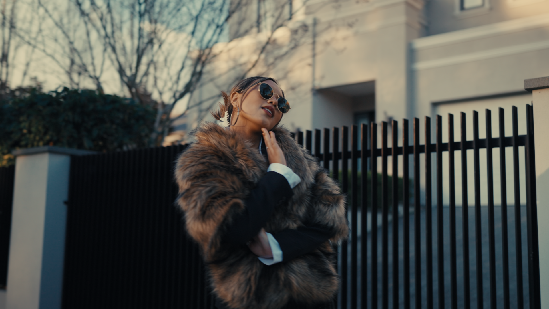 A woman with sunglasses and hoop earrings poses outdoors next to a black fence, wearing a fur coat on a chilly day with bare trees in the background.