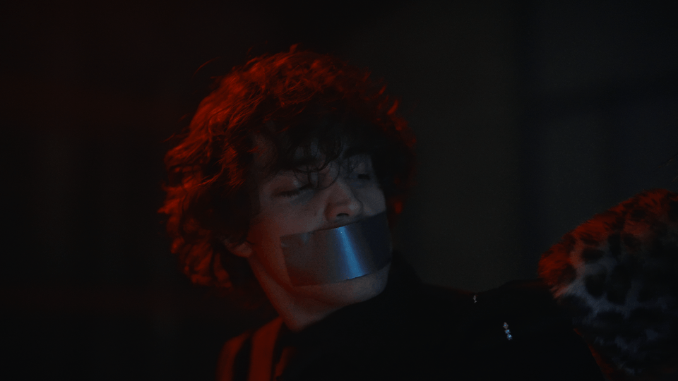 Person with curly hair and tape over mouth, in a dark setting with red lighting. Music video with Melbourne-based singer Alisya Rae, dark romance music video, batman, catwoman, Ariana Grande, Melbourne Music Video production company. Pop R&B.