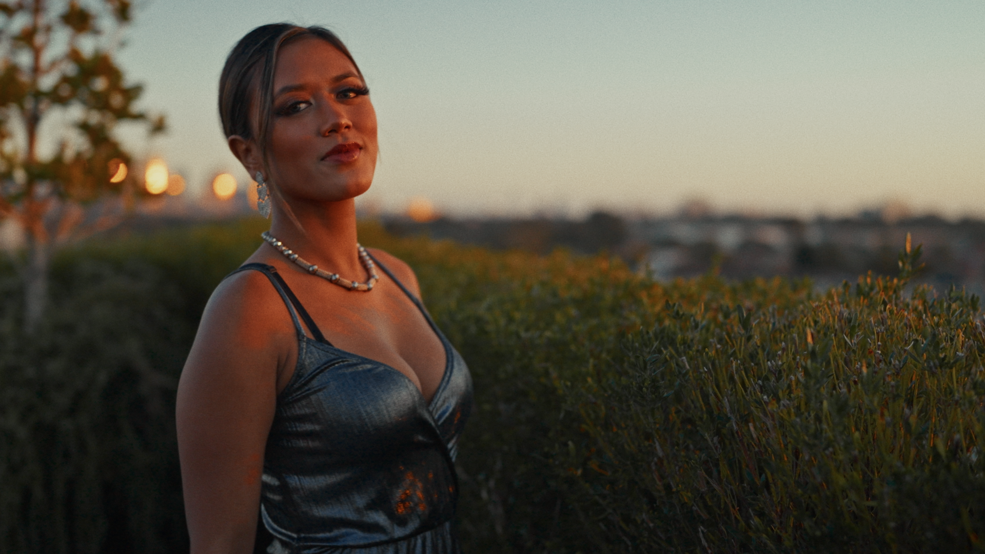 A woman standing outdoors during sunset, wearing a dark metallic dress and jewelry, with greenery and a distant horizon in the background.
