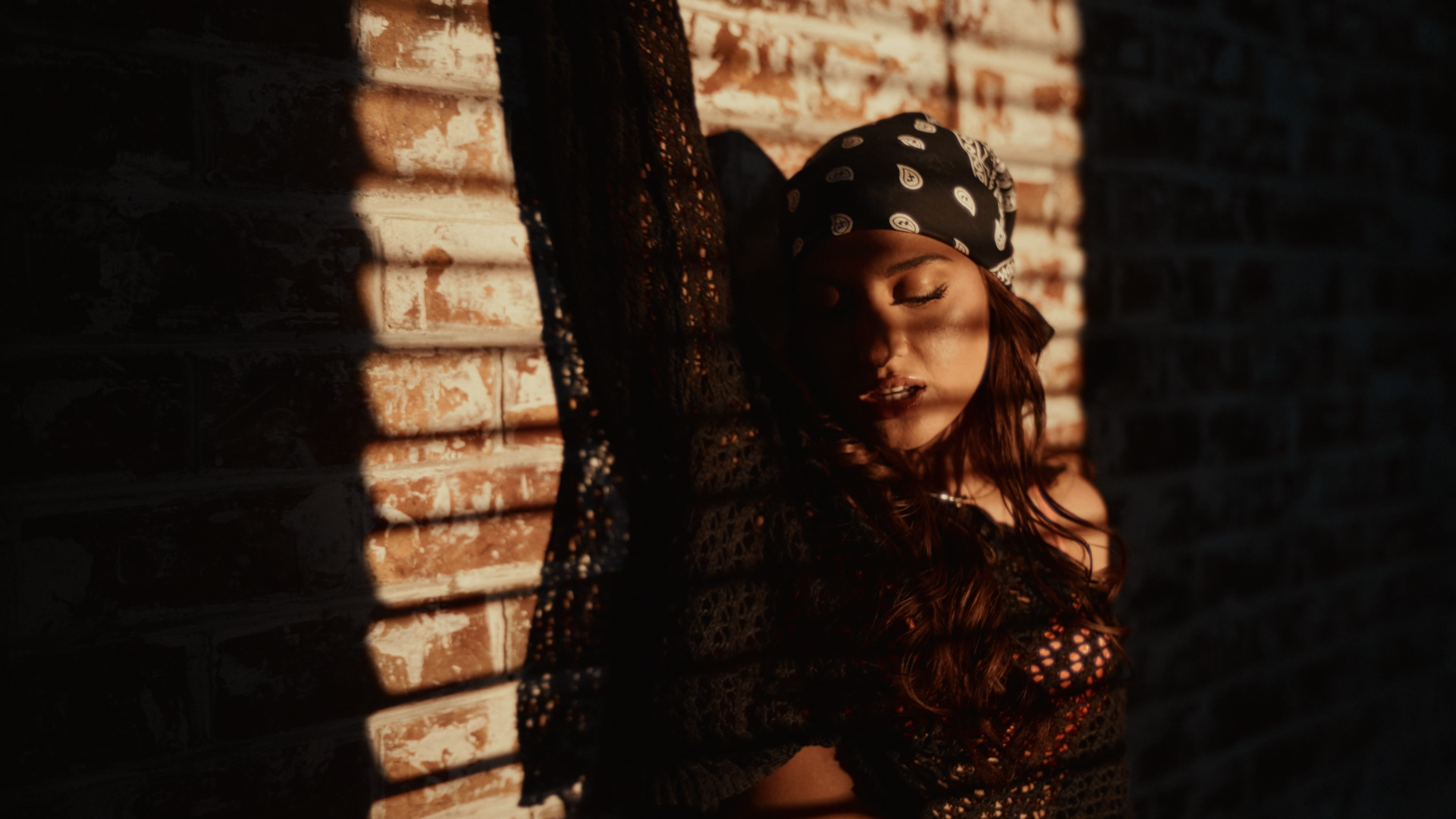 A woman with long, wavy hair wearing a patterned bandana, leaning against a brick wall with sunlight creating shadows. Music Video, Pop music video, Melbourne-based pop music artist Aanya. Melbourne music video, dance pop. Player music video.