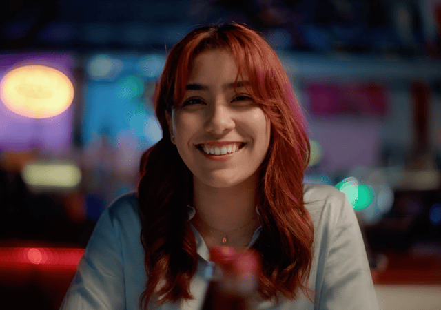 A young woman with red hair smiling in a dimly lit, vibrant setting, possibly a bar or city street at night.