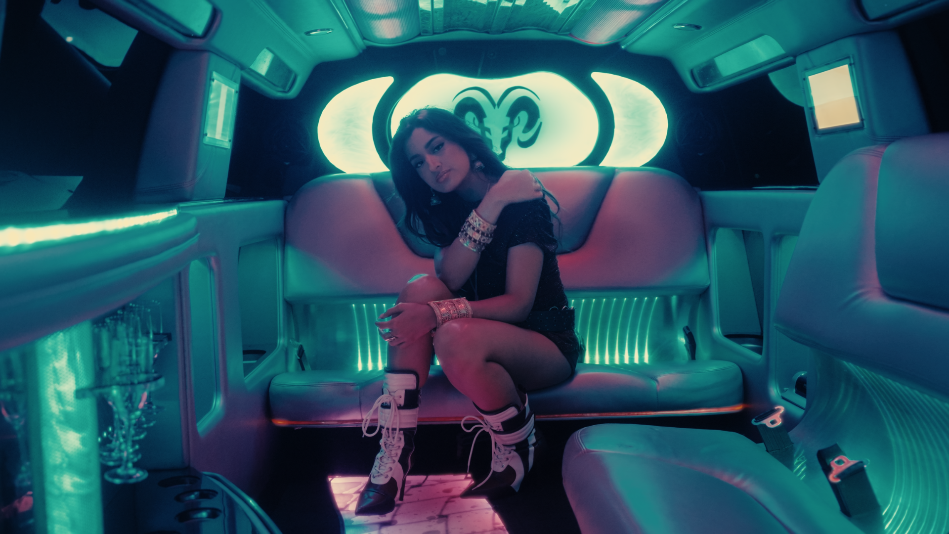 A woman sitting inside a neon-lit limousine with white leather seats, wearing a black dress, sneakers, and jewelry, with a neon sign behind her. Take Me Away music video Aanya Melbourne Music Video.