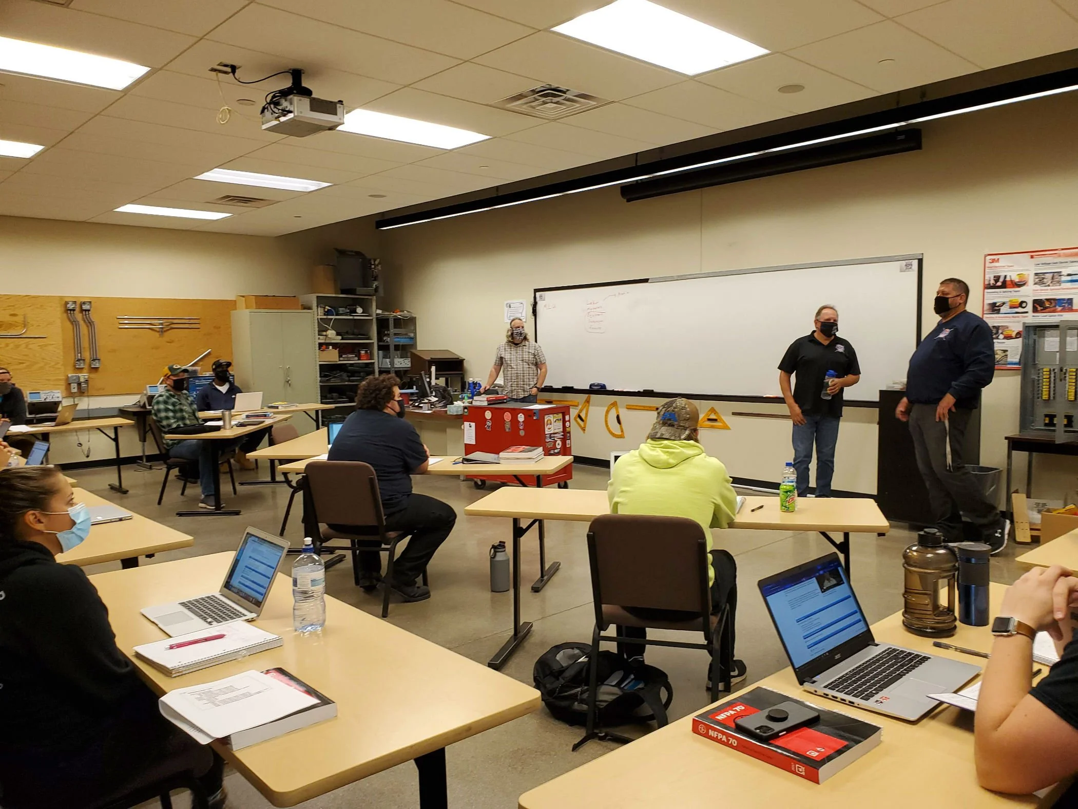A classroom setting with multiple students wearing face masks, some using laptops, and a few instructors standing and engaging with the group. The room has tables, a whiteboard, and technical equipment.