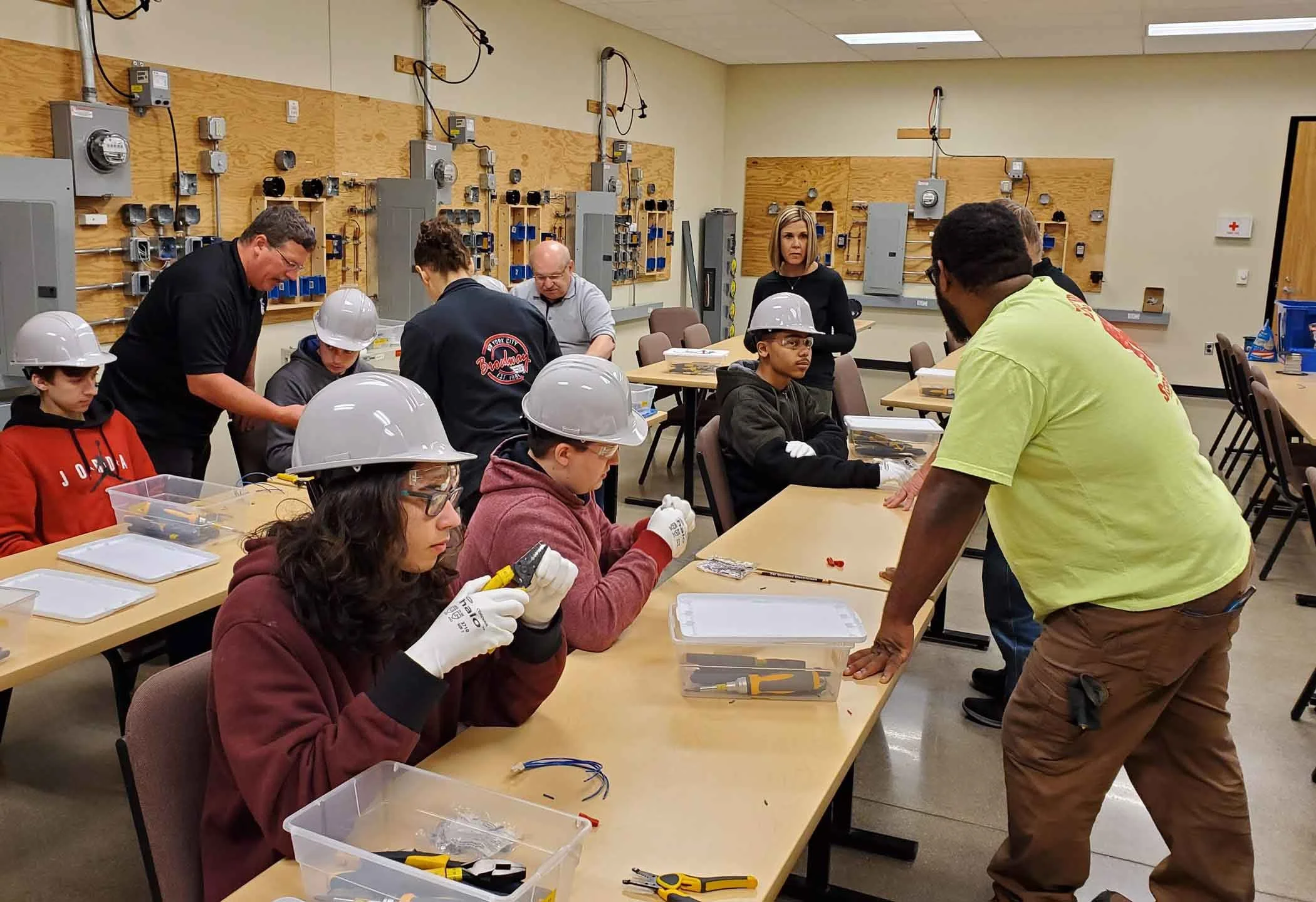 People in a classroom working with electrical components, surrounded by tools and electrical panels on the walls, some wearing safety helmets and gloves.
