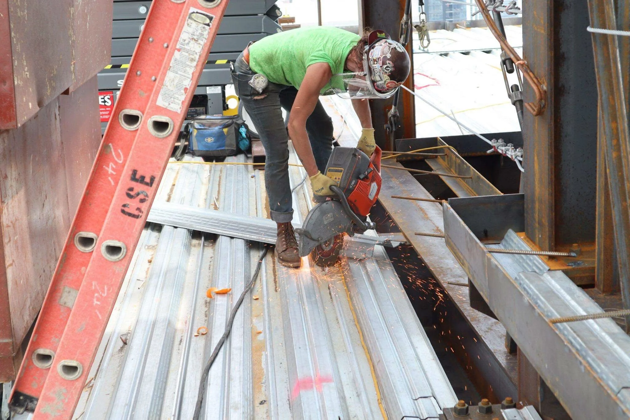 A construction worker wearing a green shirt, yellow gloves, a helmet, and safety glasses is cutting metal with a power saw on a construction site. Sparks are flying from the cutting process, and there is a ladder and various tools around.