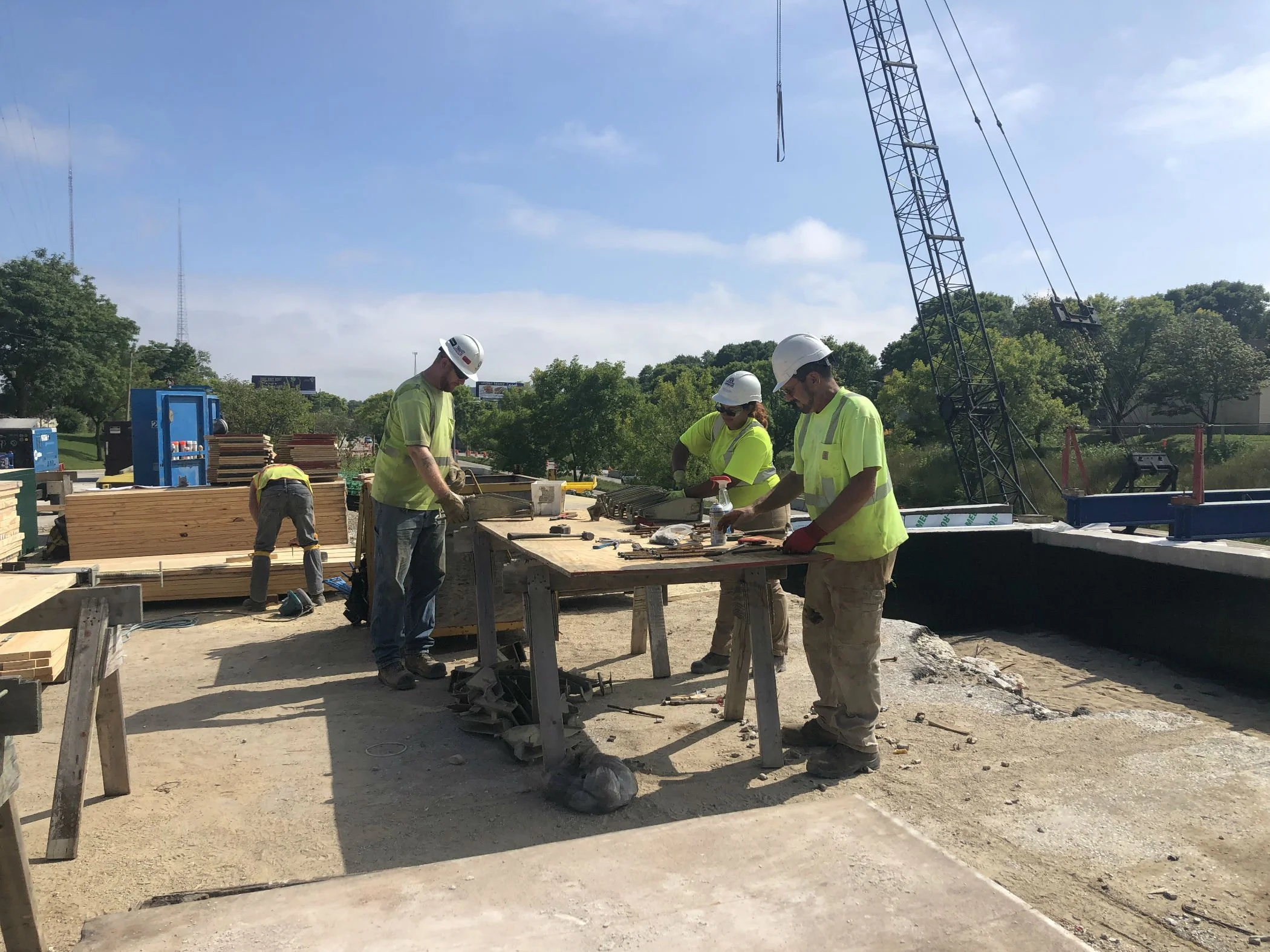 Construction workers wearing white hard hats and yellow safety vests working on a wooden platform at a construction site under a blue sky with some clouds. A crane is visible in the background, along with trees and other construction equipment.